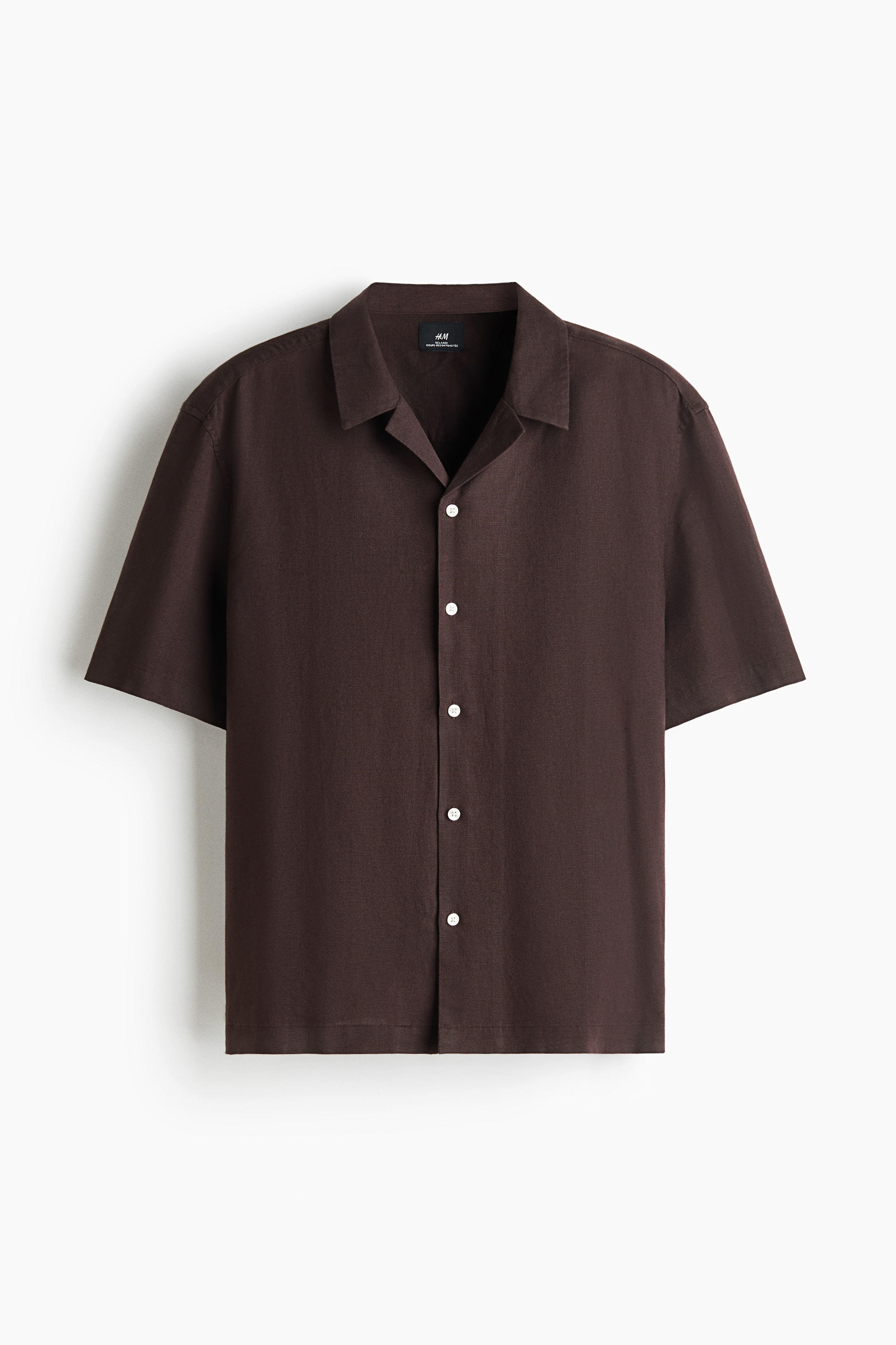Ngưá»i mẫu Äang mặc má»t chiếc áo sÆ¡ mi ngắn tay mà u nâu sẫm, vá»i chất liá»u dá»t cotton-linen có bá» mặt sần. Ão có phom dáng thoải mái, cá» resort, nẹp áo kiá»u French front vá»i cúc trắng và gấu áo thẳng, ÄÆ°á»£c phá»i cùng quần á»ng rá»ng mà u kem.