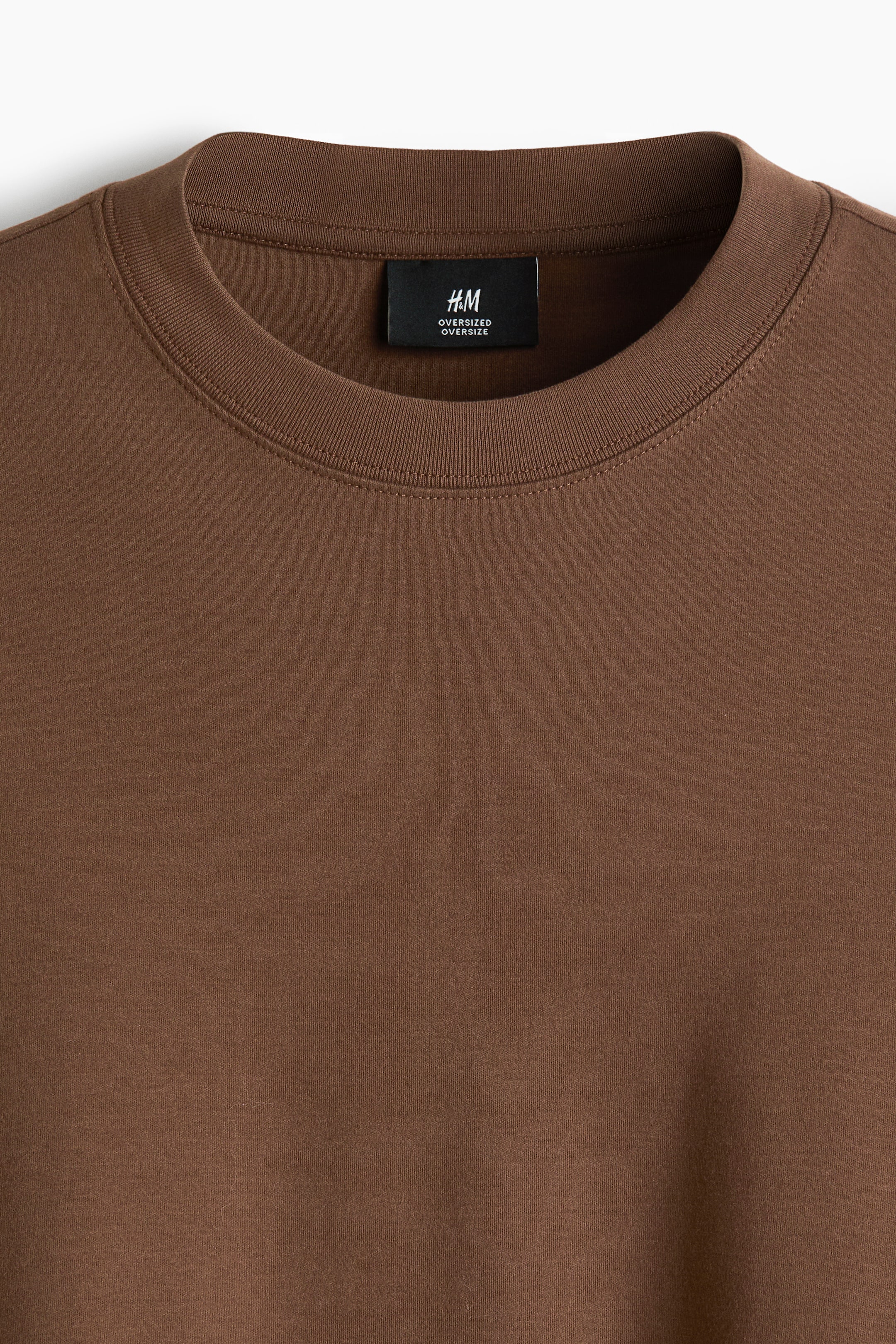 Visualizza immagine più grande: Una t-shirt marrone viene presentata in una vista frontale, con un collo a coste. Un'etichetta del marchio nera, contrassegnata con il testo "H&M OVERSIZED", è visibile sulla parte interna posteriore del collo.