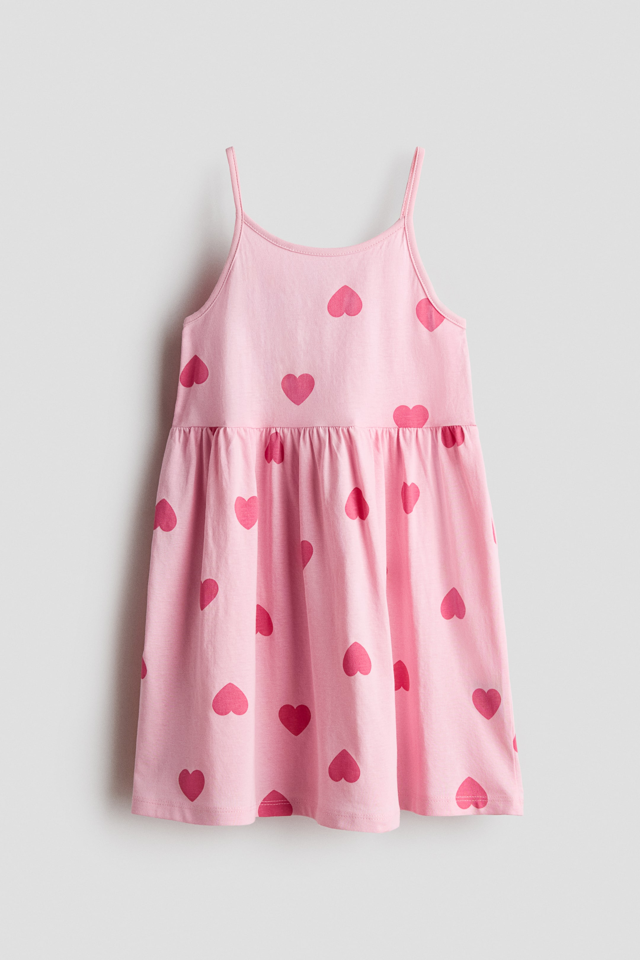 View larger image: A dress crafted from soft cotton jersey, featuring slender shoulder straps, a gathered seam at the waist, and a subtly flared skirt, is showcased in its front view. The dress is adorned with a pink hue patterned with darker pink hearts.