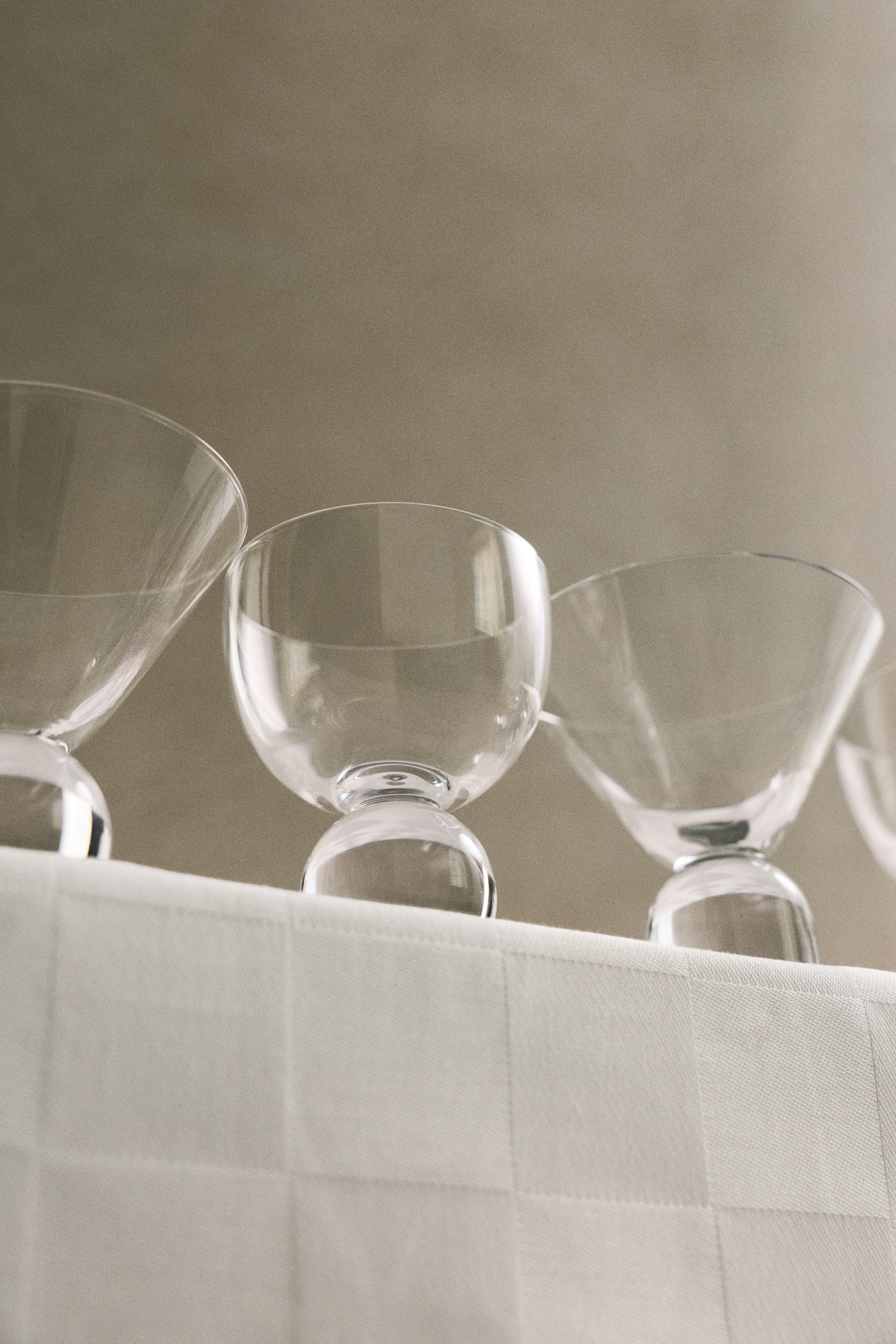 Grotere afbeelding bekijken: Helder glazen cocktailglazen staan in een rij op een wit geruit tafelkleed, waarbij elk glas een uniek, sculpturaal ontwerp heeft. De glazen vertonen verschillende kelkvormen, waaronder conische en afgeronde silhouetten, ondersteund door dikke, massieve glazen voetstukken in bol- en cilindervorm.