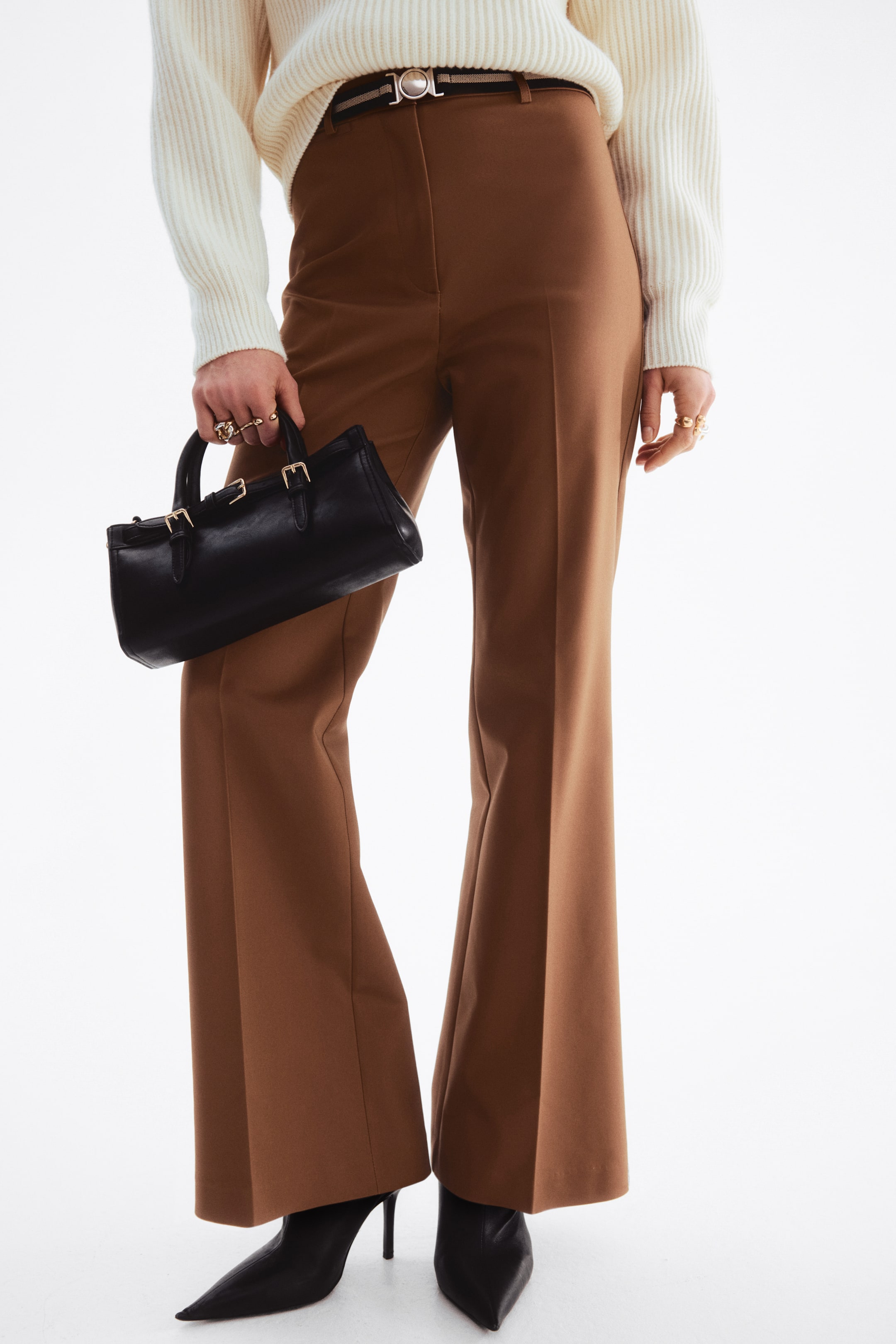 View larger image: Brown tailored trousers boast a high-waisted design and a unique flare from the knee down, with the hems elegantly draping over the tops of pointed-toe boots.