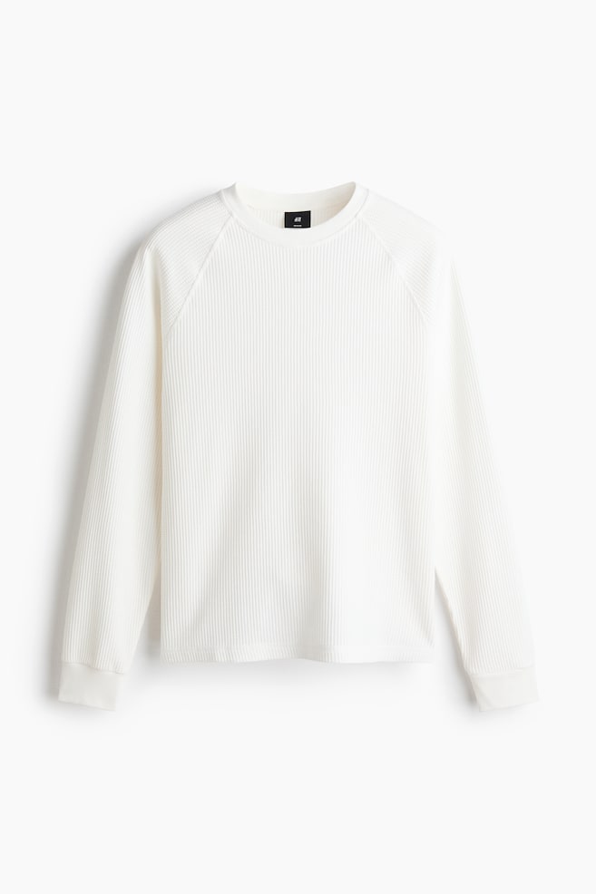 A white long-sleeved waffle knit shirt is displaying its front view, featuring a crew neck, raglan sleeves, and ribbed cuffs. - 1