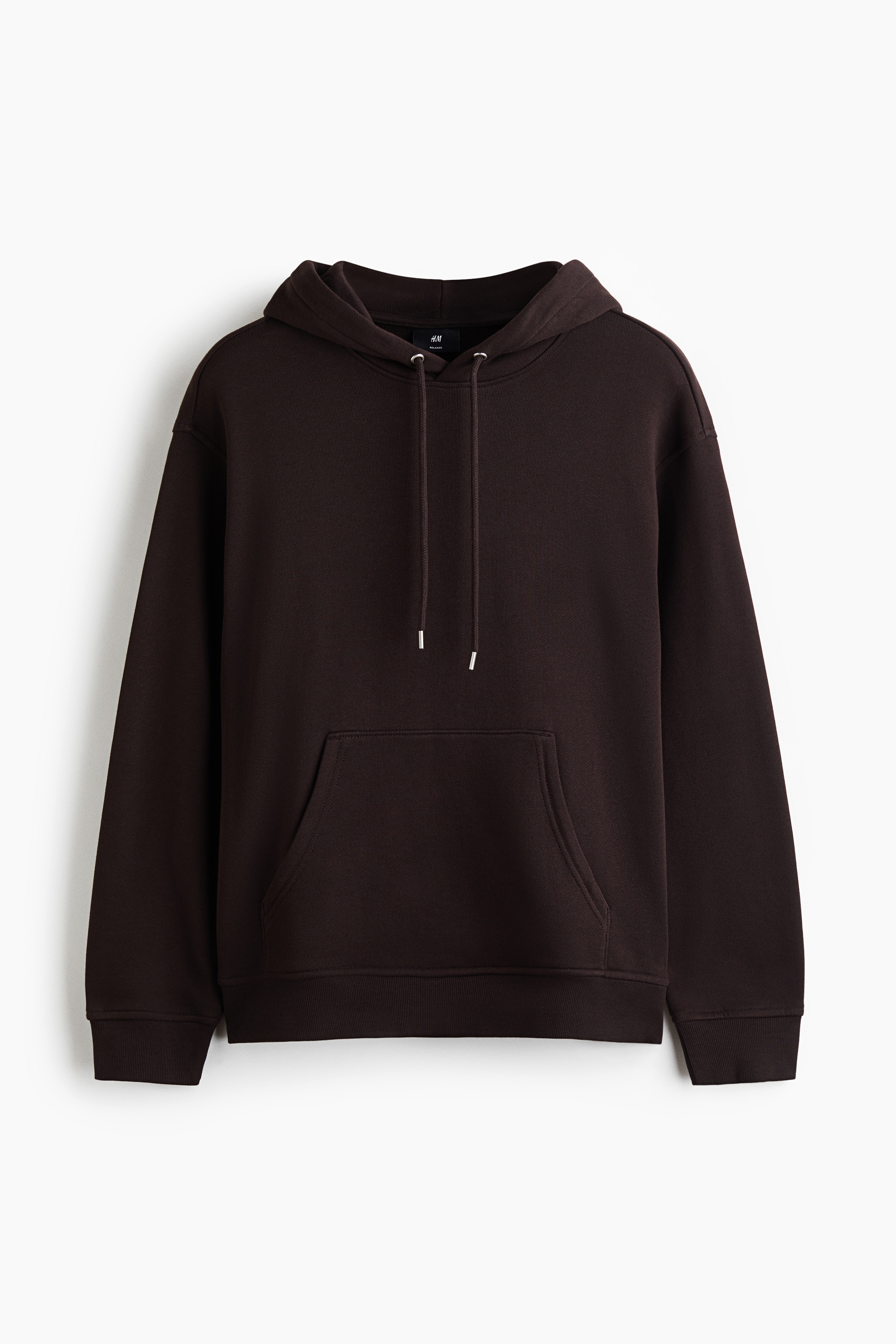 A dark brown cotton sweatshirt hoodie is being worn by the model, featuring a relaxed fit, a drawstring hood, dropped shoulders, and a front kangaroo pocket. The hoodie is paired with black trousers, with the model standing with one hand resting in the trouser pocket.