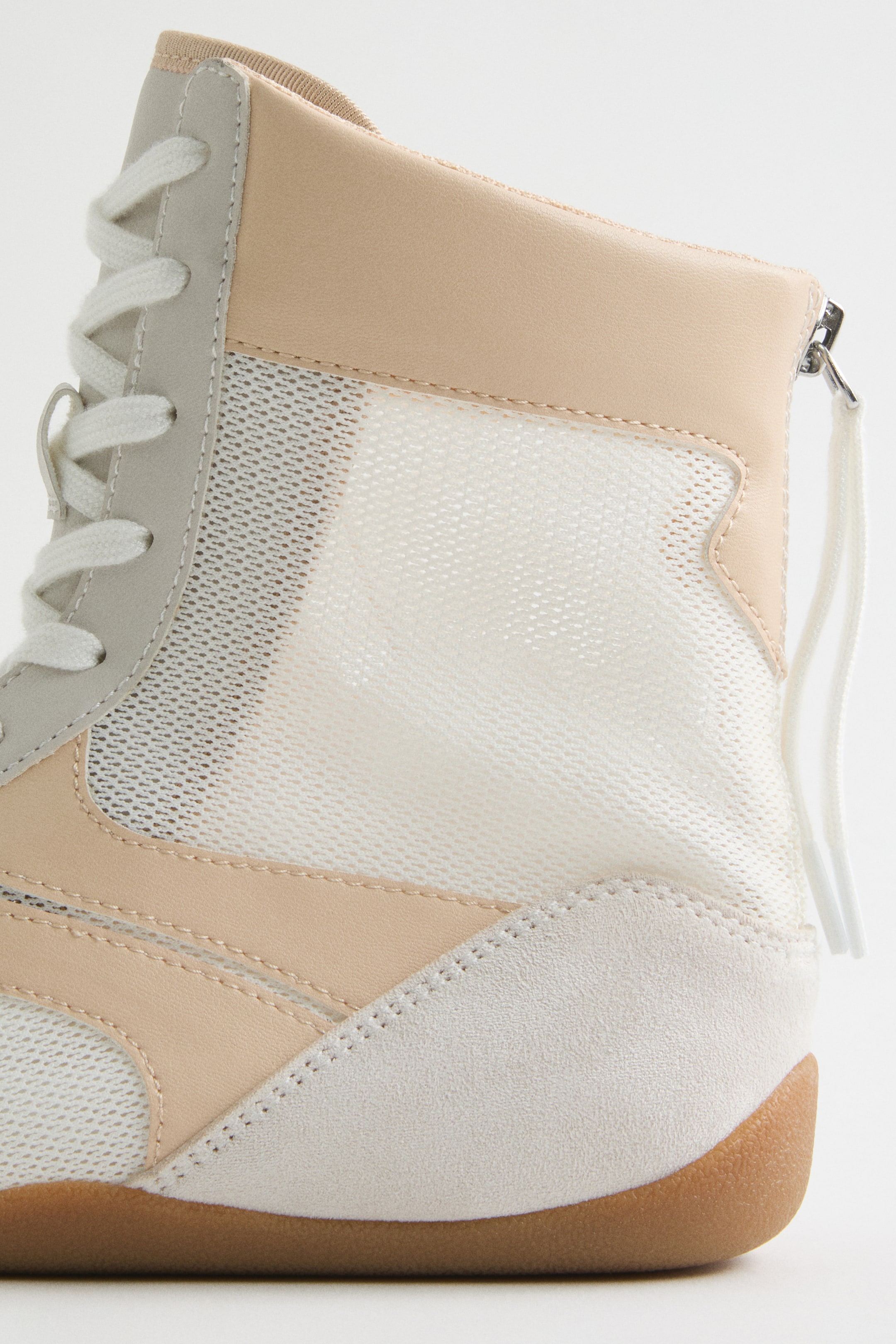 Größeres Bild ansehen: Ein High-Top-Sneaker mit hellbeigem Leder und weißen Mesh-Einsätzen an der Oberseite, akzentuiert mit hellgrauem Wildleder-ähnlichem Material an Ferse und Zehen, aus einem seitlich-hinteren Winkel betrachtet. Weiße Schnürsenkel verlaufen an der Seite nach oben, und ein silberner Reißverschlussanhänger mit weißer Schnur ist am Rücken sichtbar, wobei der Schuh auf einer braunen Gummisohle ruht.