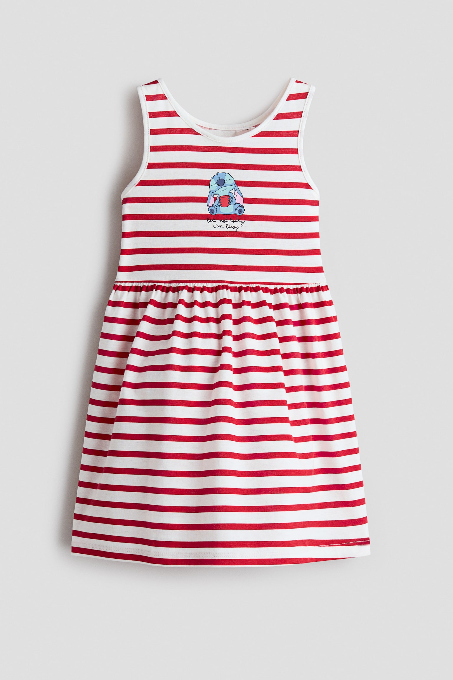 A sleeveless cotton jersey dress is shown from the front, featuring an all-over pattern of red and white horizontal stripes. The dress displays a round neckline, a gathered seam at the waist that creates a flared skirt, and a small cartoon graphic at the center chest.