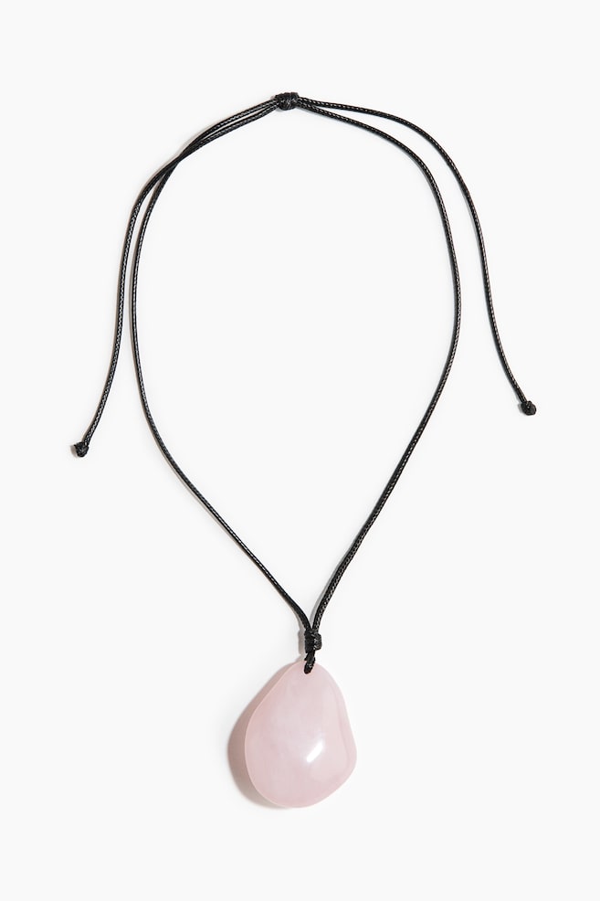 A necklace is lying flat, showcasing a light pink, irregularly shaped stone pendant. It is suspended from a black cord featuring an adjustable sliding knot at the top. - 1