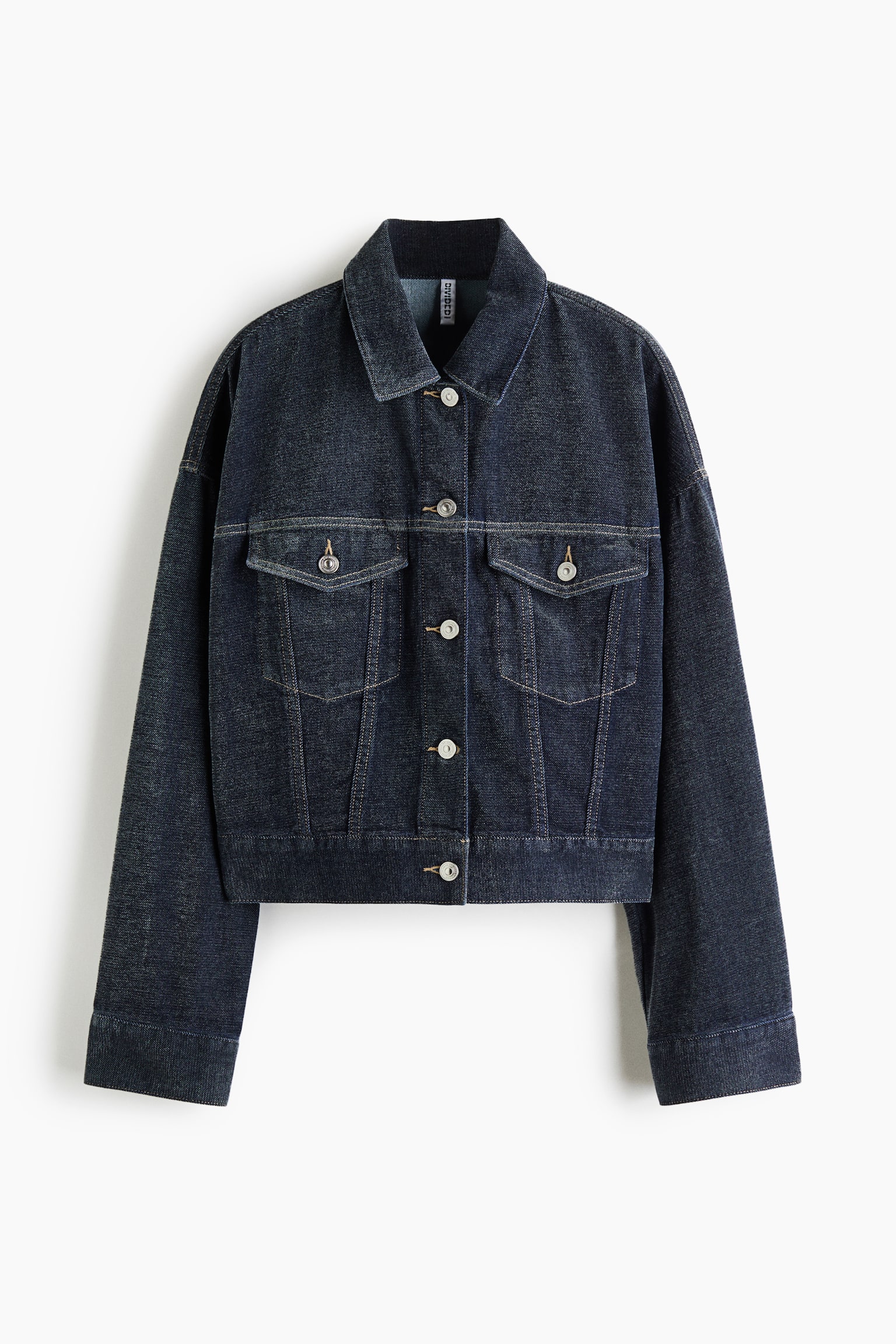 A dark-wash denim jacket is shown from the front, featuring a spread collar, dropped shoulders, and a button-front fastening with silver-tone buttons. The jacket features two button-flap chest pockets, subtle contrast stitching, and a slightly cropped, straight hem.