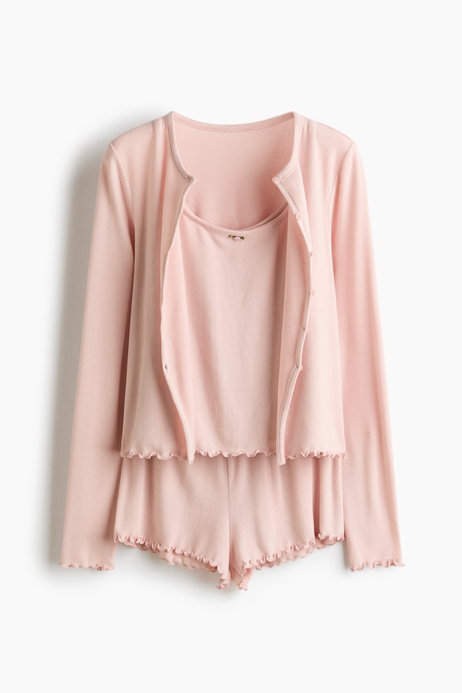 A light pink three-piece pyjama set is hanging in a front view, featuring a long-sleeved cardigan worn open over a matching scoop-neck camisole and shorts. All three pieces are showcasing lettuce-edge trims on the hems and cuffs, with a small decorative bow detailing the center of the camisole's neckline. - 1