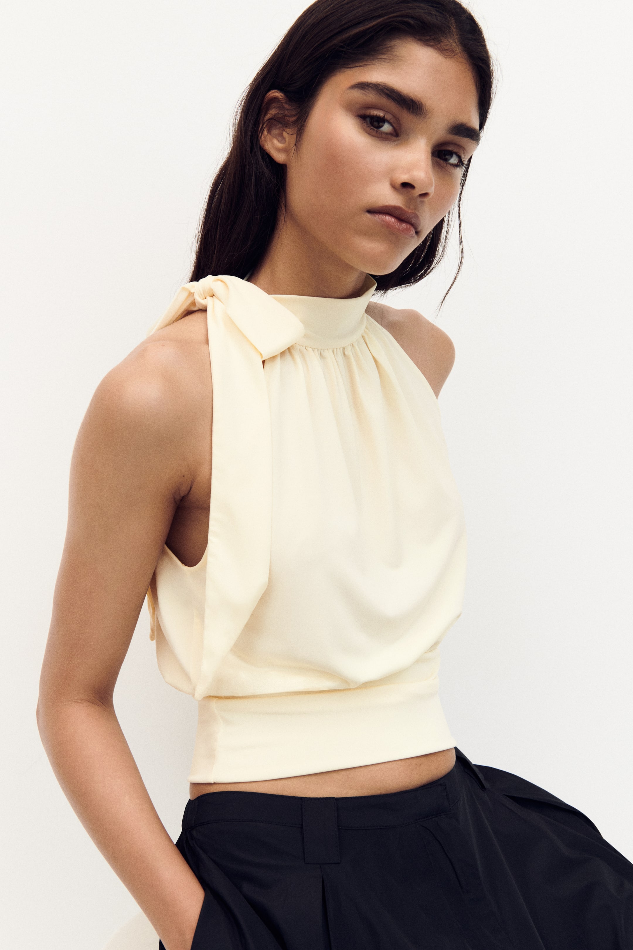 View larger image: A light yellow halter neck top features a high, gathered neckline, with self-tie shoulder straps forming a bow on the left shoulder. The top concludes with a fitted, cropped hem.