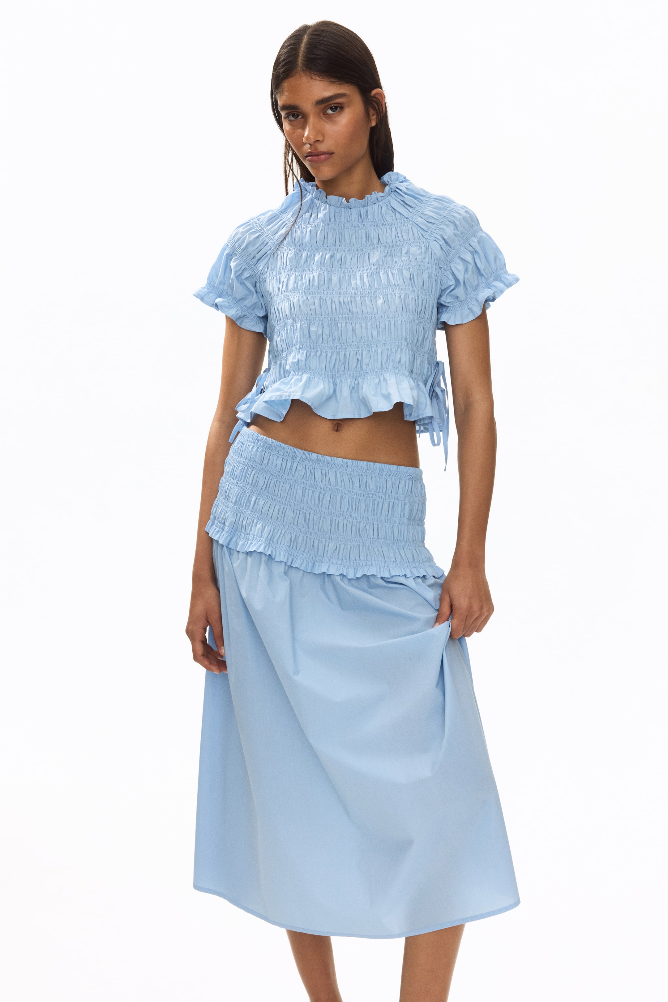 View larger image: The model is showcasing a light blue, short, fitted blouse, meticulously crafted from a smocked cotton weave. This blouse is adorned with frill trims along the round neckline, short raglan sleeves, and hem, with ties accentuating the side slits at the hem. Complementing the blouse, the model is also wearing a matching smocked skirt.