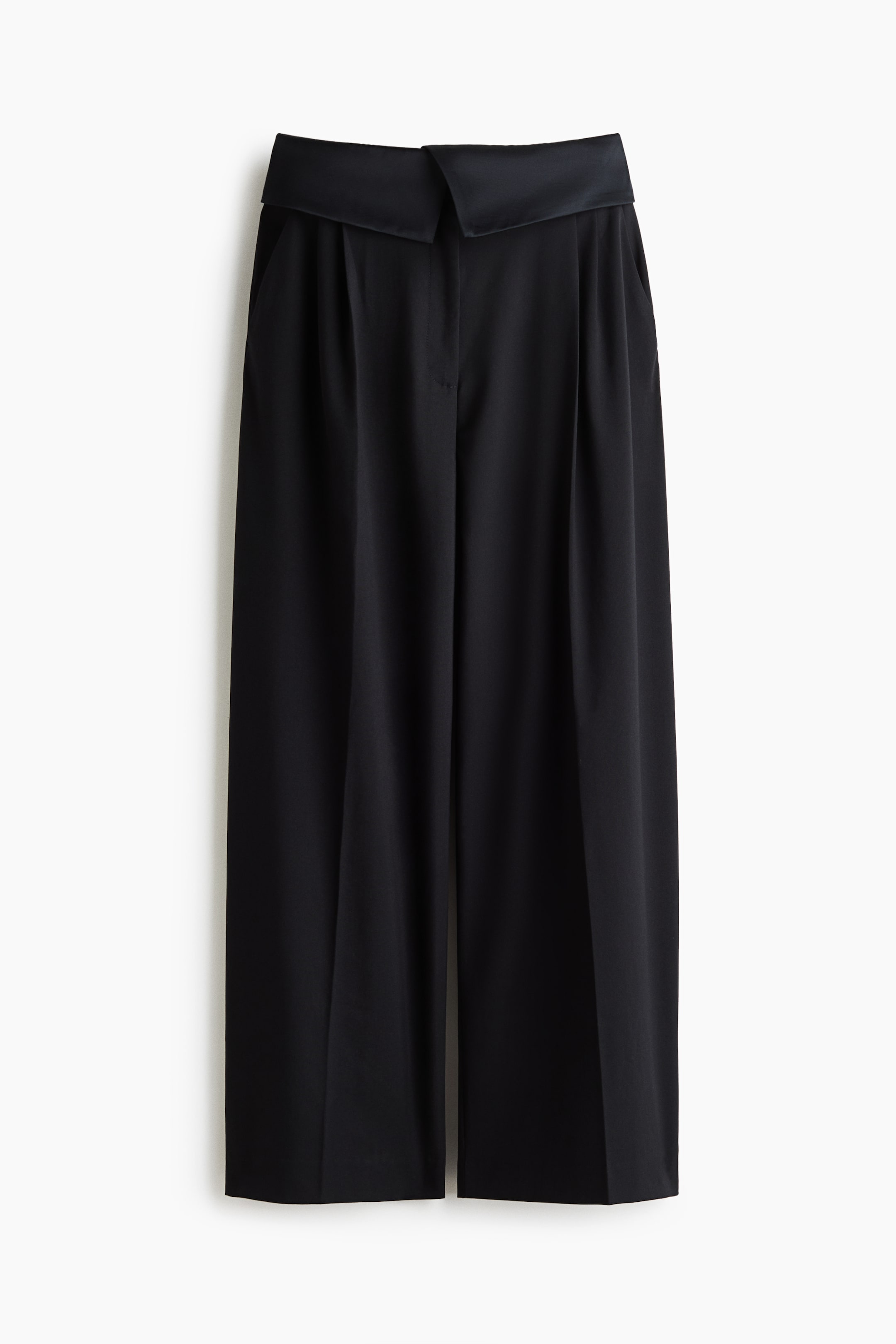 View larger image: Black loose-fit trousers in a woven fabric are shown from the front, showcasing a high waist with a wide, overlapping foldover waistband. The trousers feature front pleats, subtle diagonal side pockets, and wide legs with sharp creases running down the centre.