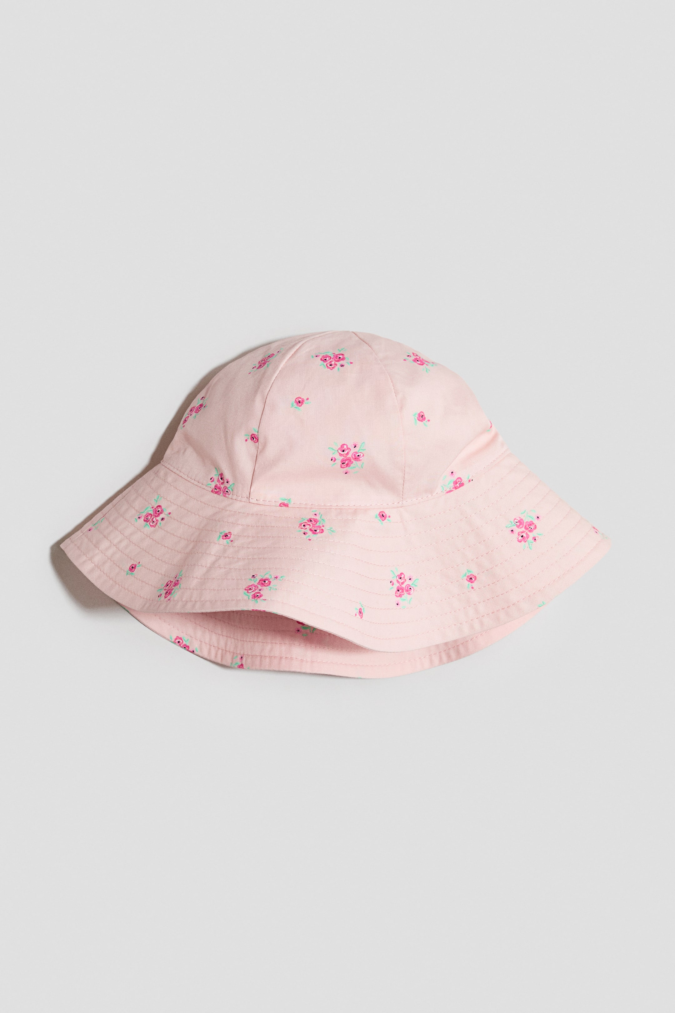 View larger image: A sun hat is presented from a front view, crafted from a light pink soft cotton weave. It features an all-over floral print of small pink roses and a wide brim with visible stitching lines.