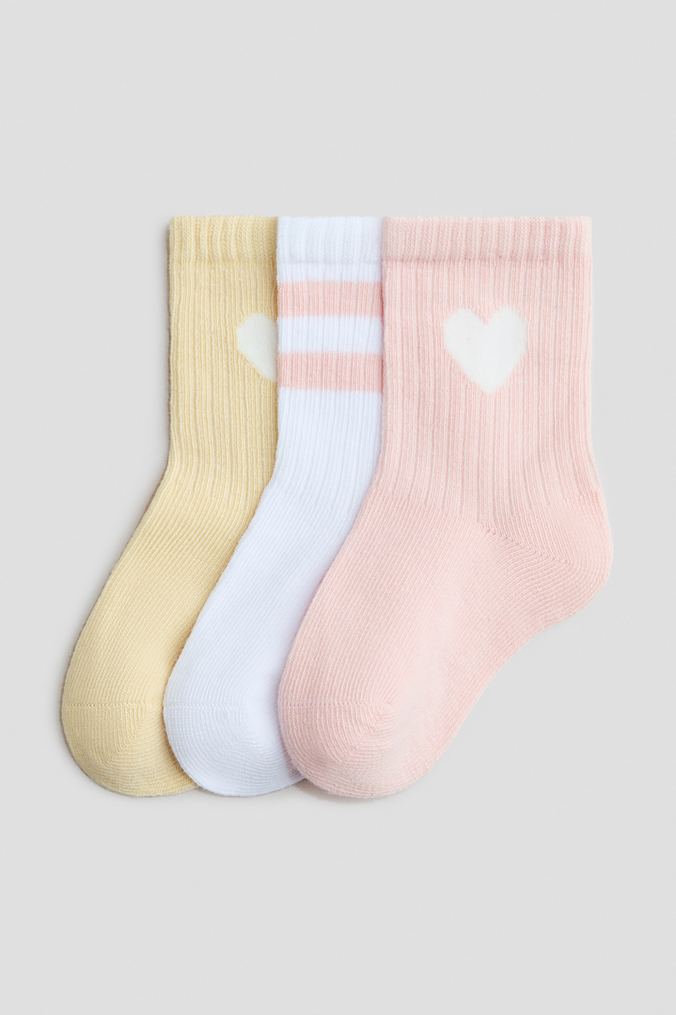 View larger image: A set of three fine-knit socks is overlapping, each featuring a ribbed cuff. The leftmost sock is yellow with a white heart motif, the middle sock is white with two light pink stripes, and the rightmost sock is light pink with a white heart motif.