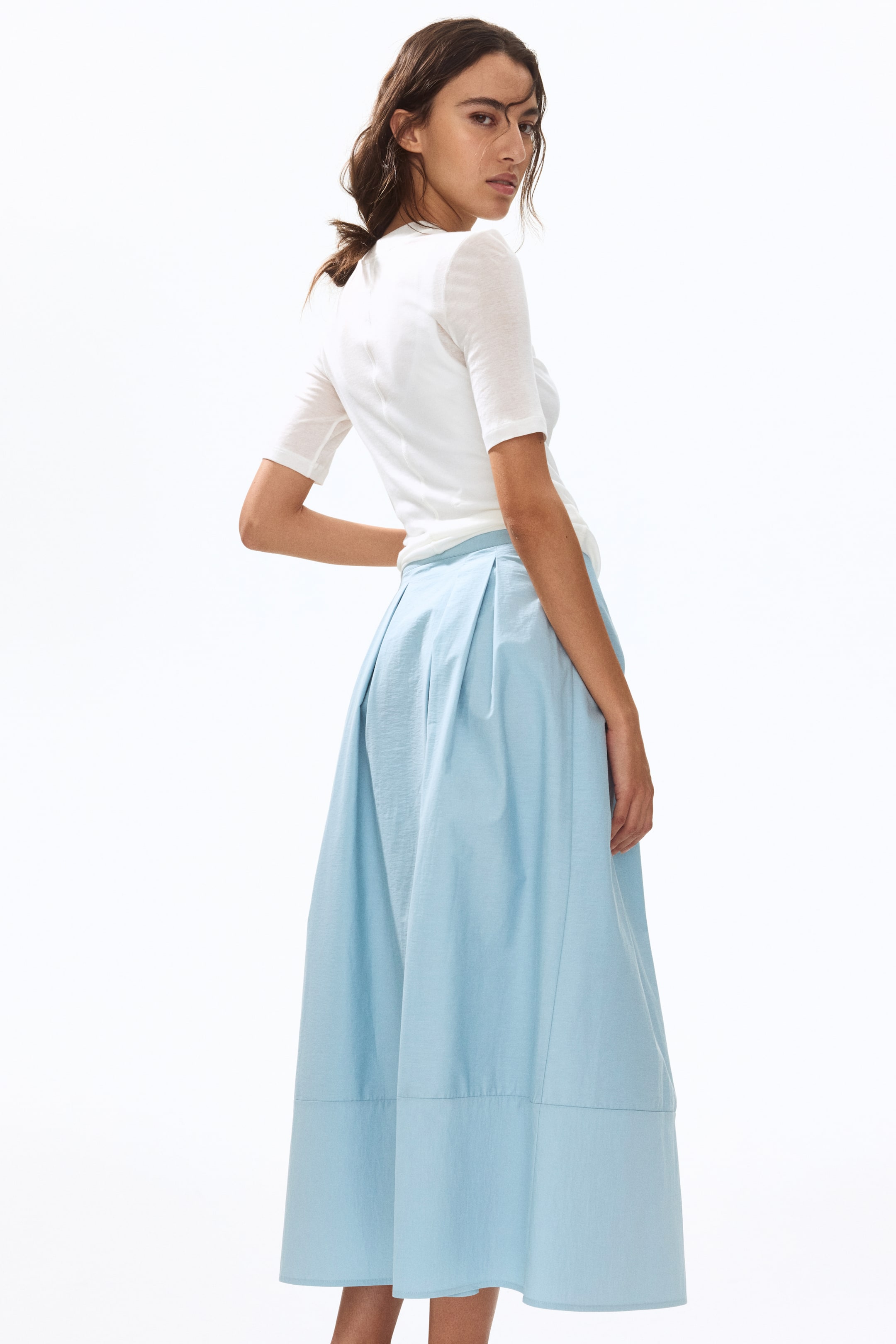View larger image: The model is wearing a light blue calf-length A-line skirt crafted in a crisp weave. It highlights a double-layered waistband and inverted box pleats at the back. The model's white top is partially tucked into the skirt.