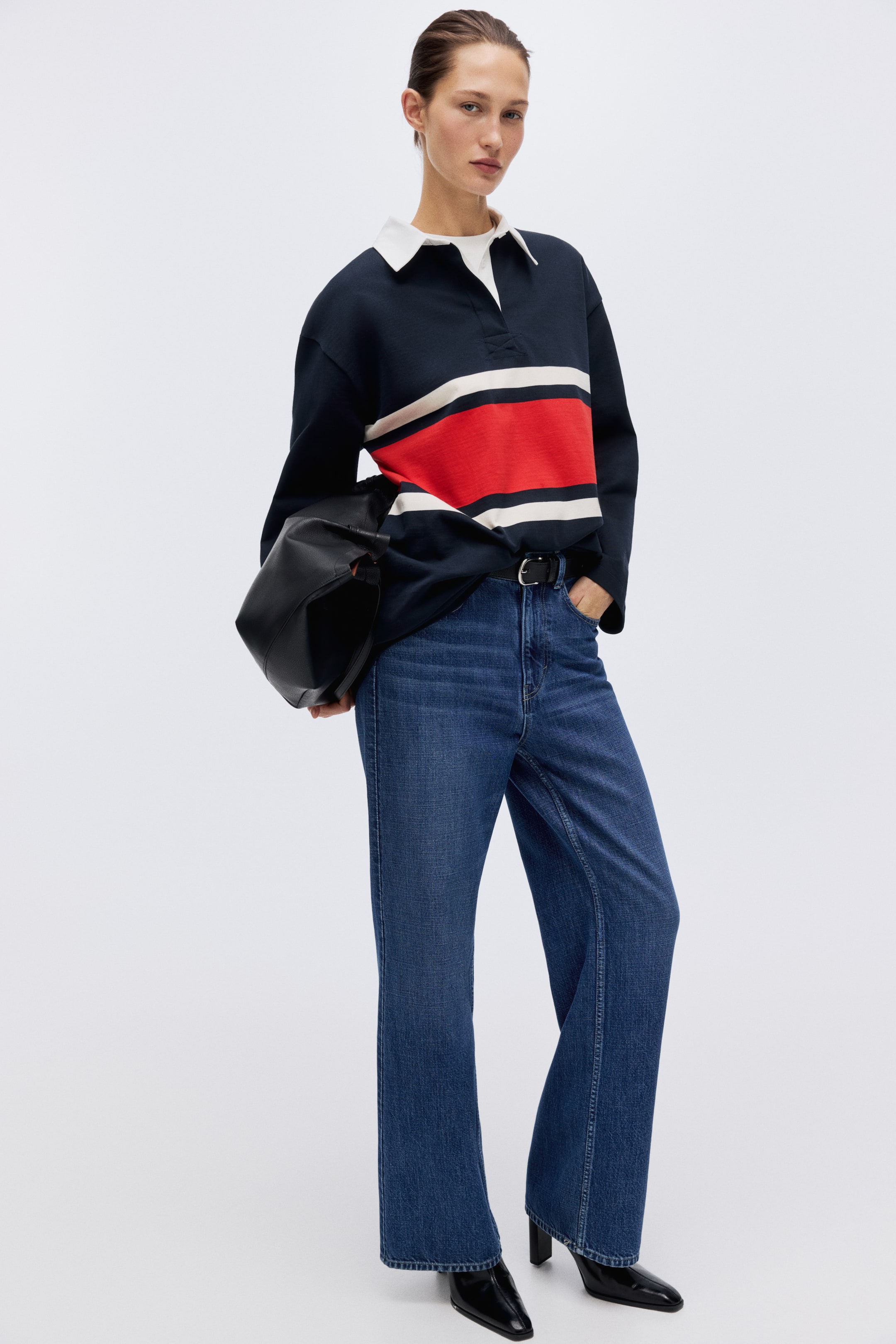 View larger image: A navy-blue long-sleeved rugby shirt features a white collar and a wide red stripe flanked by two thinner white stripes across the chest. It is styled with a visible white undershirt collar and is partially tucked into blue jeans.