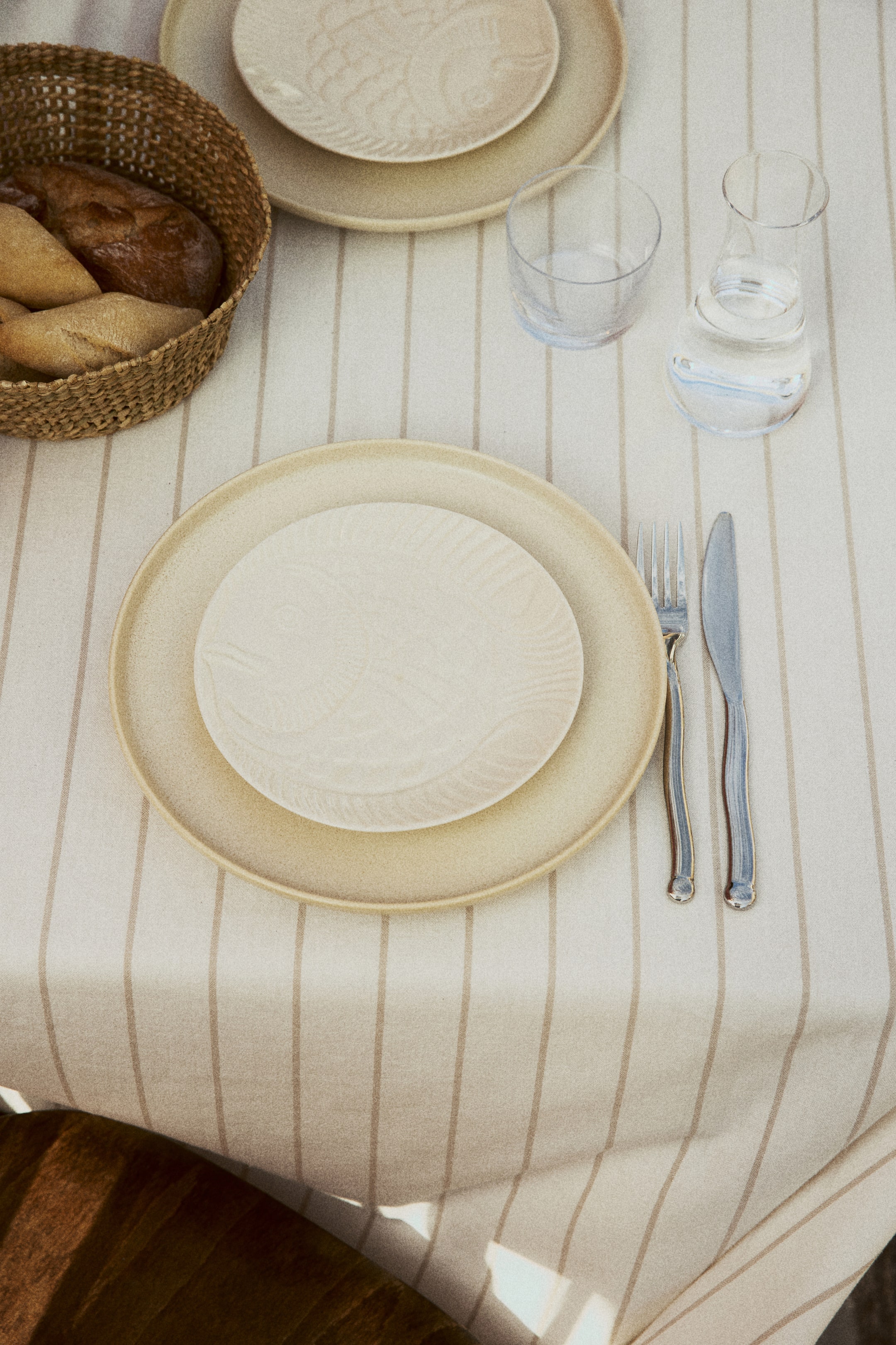 Ver imagen más grande: Un plato grande, redondo y de color beige claro está coronado por un plato más pequeño, redondo y de color blanco roto con un patrón de peces en relieve. Un tenedor y un cuchillo plateados con mangos decorativos se encuentran a la derecha de los platos.