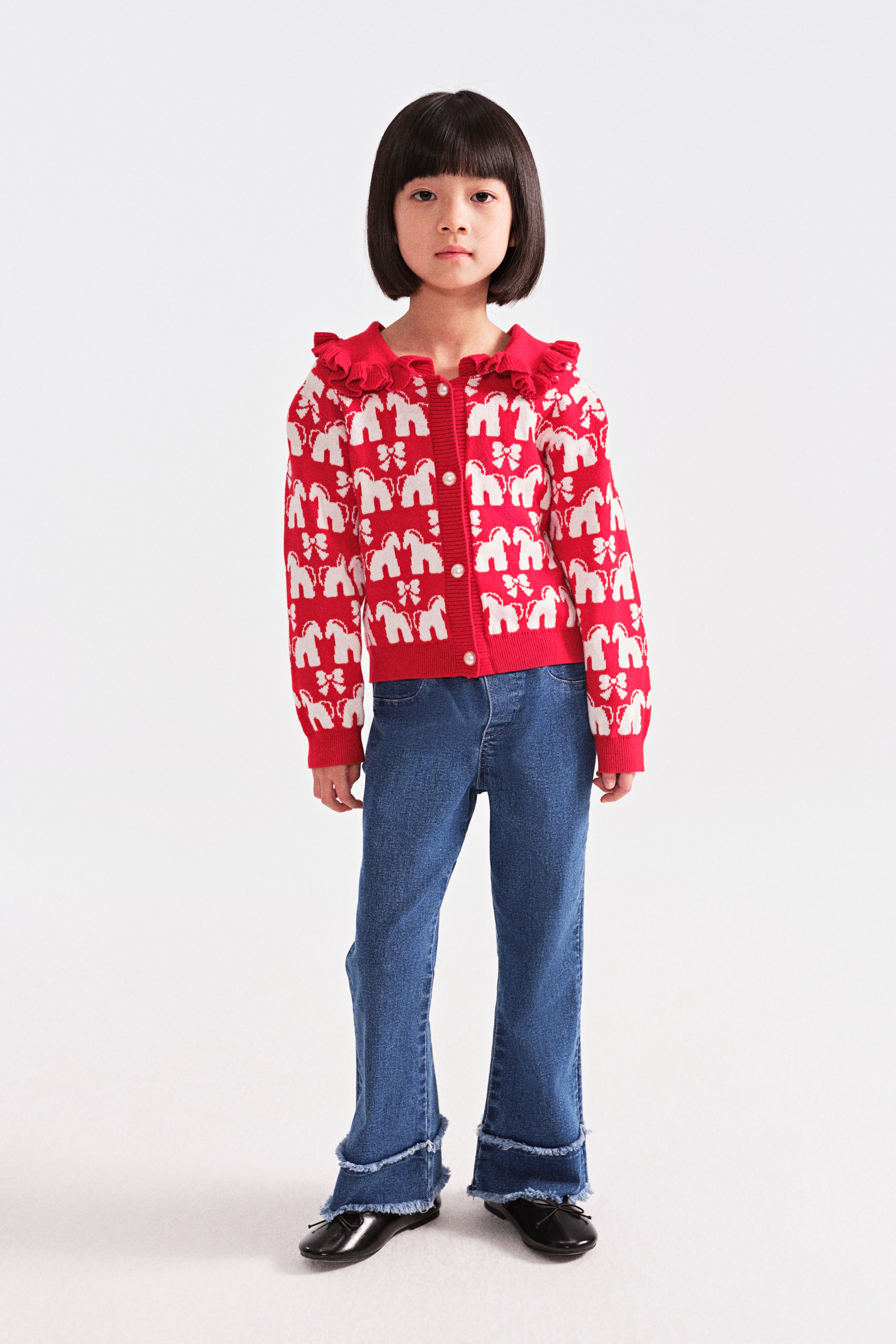 拡大画像を表示: 赤と白のニットカーディガンは、白い象とリボンの繰り返し模様が特徴です。フリル付きの赤い襟、前立てに並んだパール調のボタン、リブ編みの袖口と裾で仕上げられています。