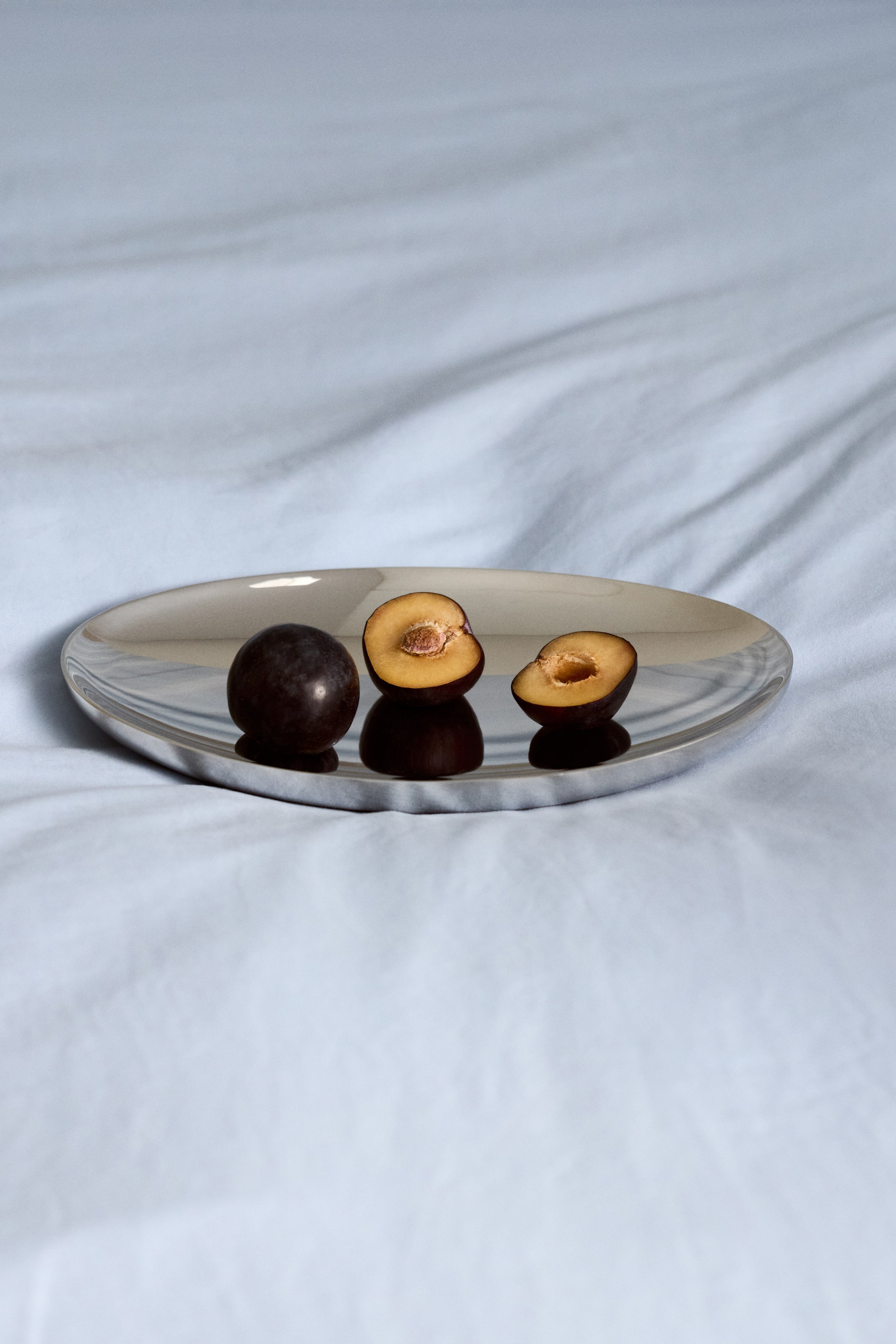 View larger image: A silver, oval-shaped reflective tray holds one whole dark plum and two plum halves. The plum halves reveal their yellow-orange flesh and pits.
