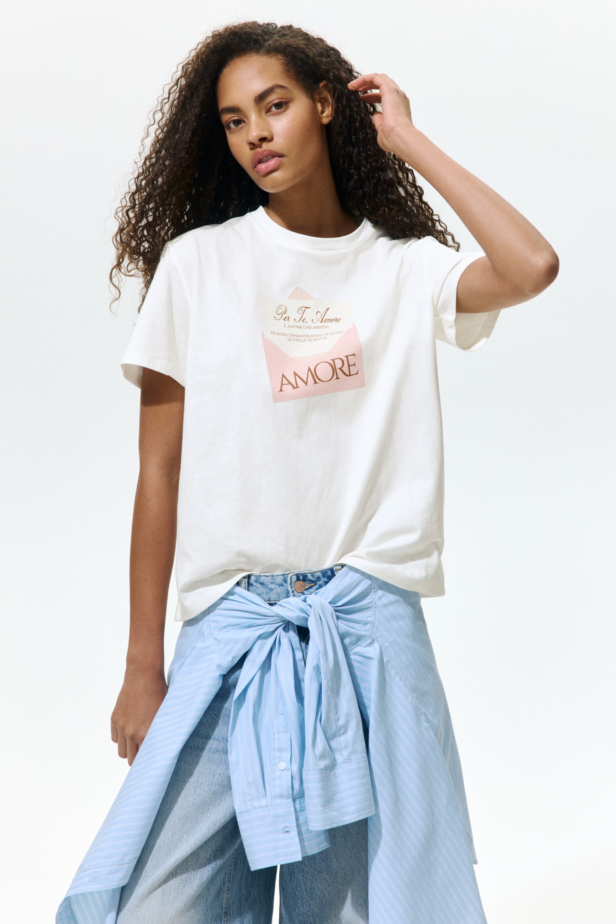 View larger image: The model is wearing a white cotton jersey T-shirt that has a round, rib-trimmed neckline, gently dropped shoulders, and a straight-cut hem. The T-shirt showcases a pink envelope graphic print with the text "AMORE" on the chest. It is styled with light-wash jeans and a light blue striped shirt tied around the waist.