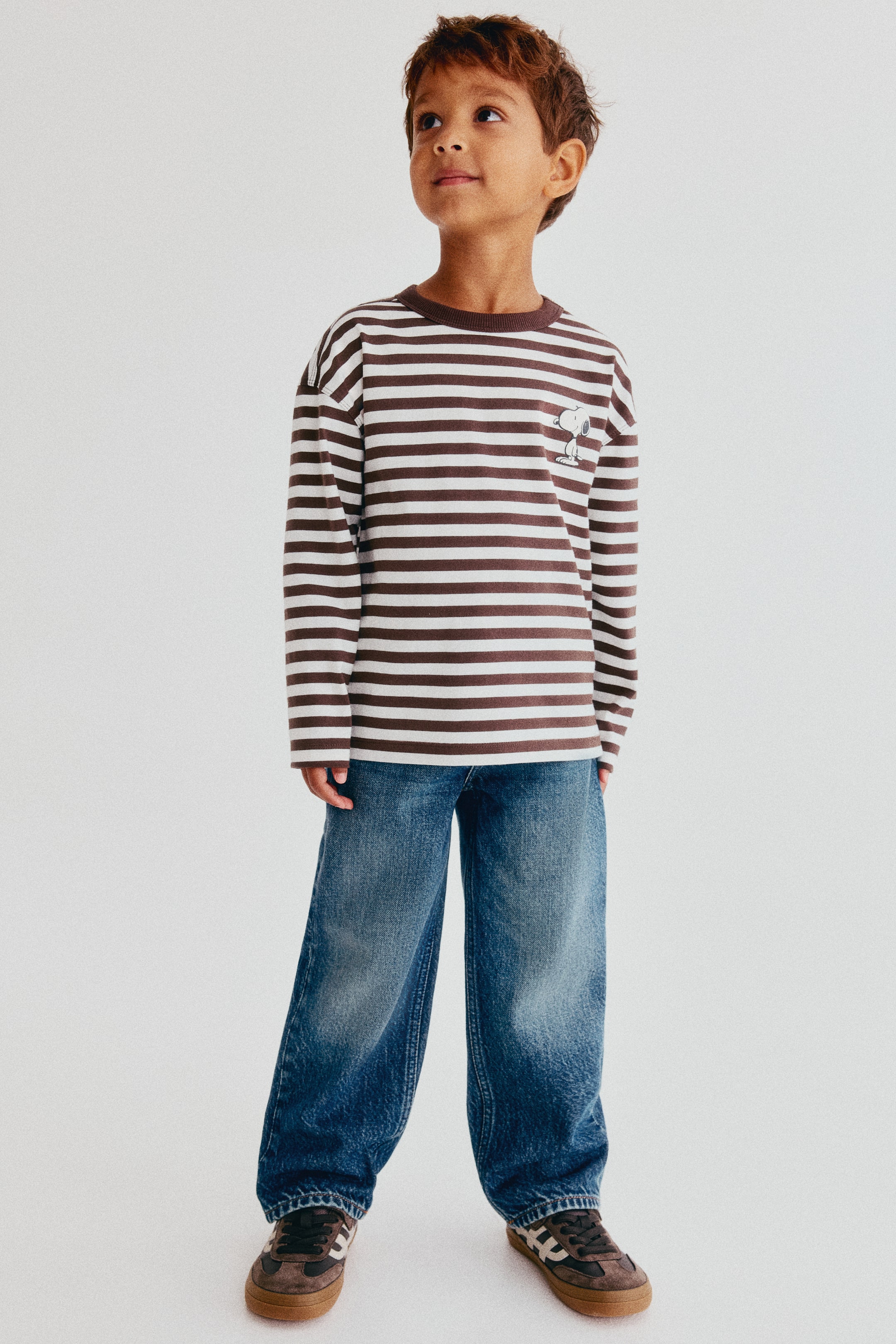 Voir une image plus grande: Ce jean en denim bleu présente une coupe ample aux hanches, aux cuisses et aux mollets, avec un délavage décoloré, vu de face. Le jean tombe librement sur des sneakers et est agencé avec une chemise à manches longues à rayures.