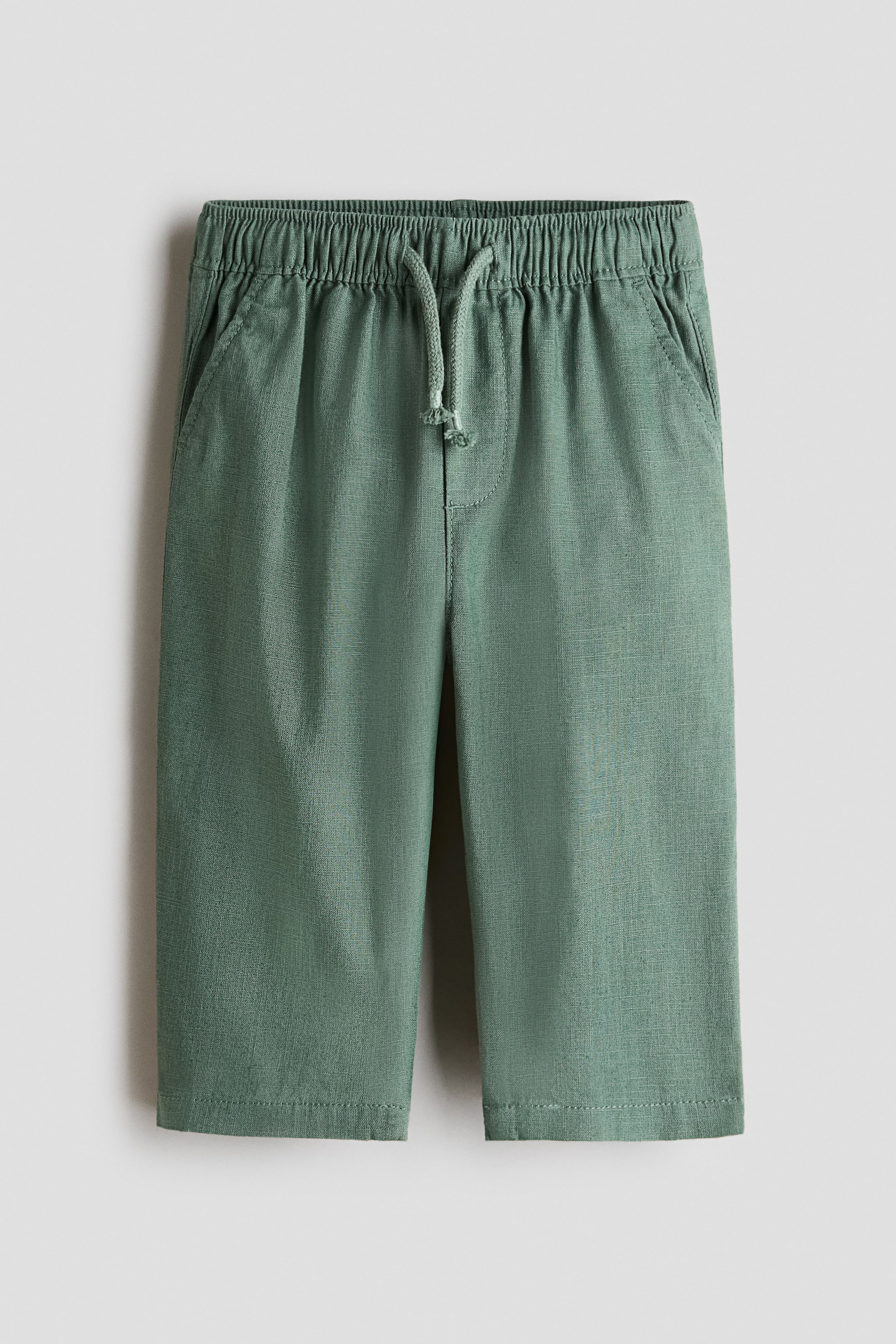 View larger image: Green pants feature an elasticated, ruched waistband with a front-tied drawstring. They have two side pockets and a visible woven texture.