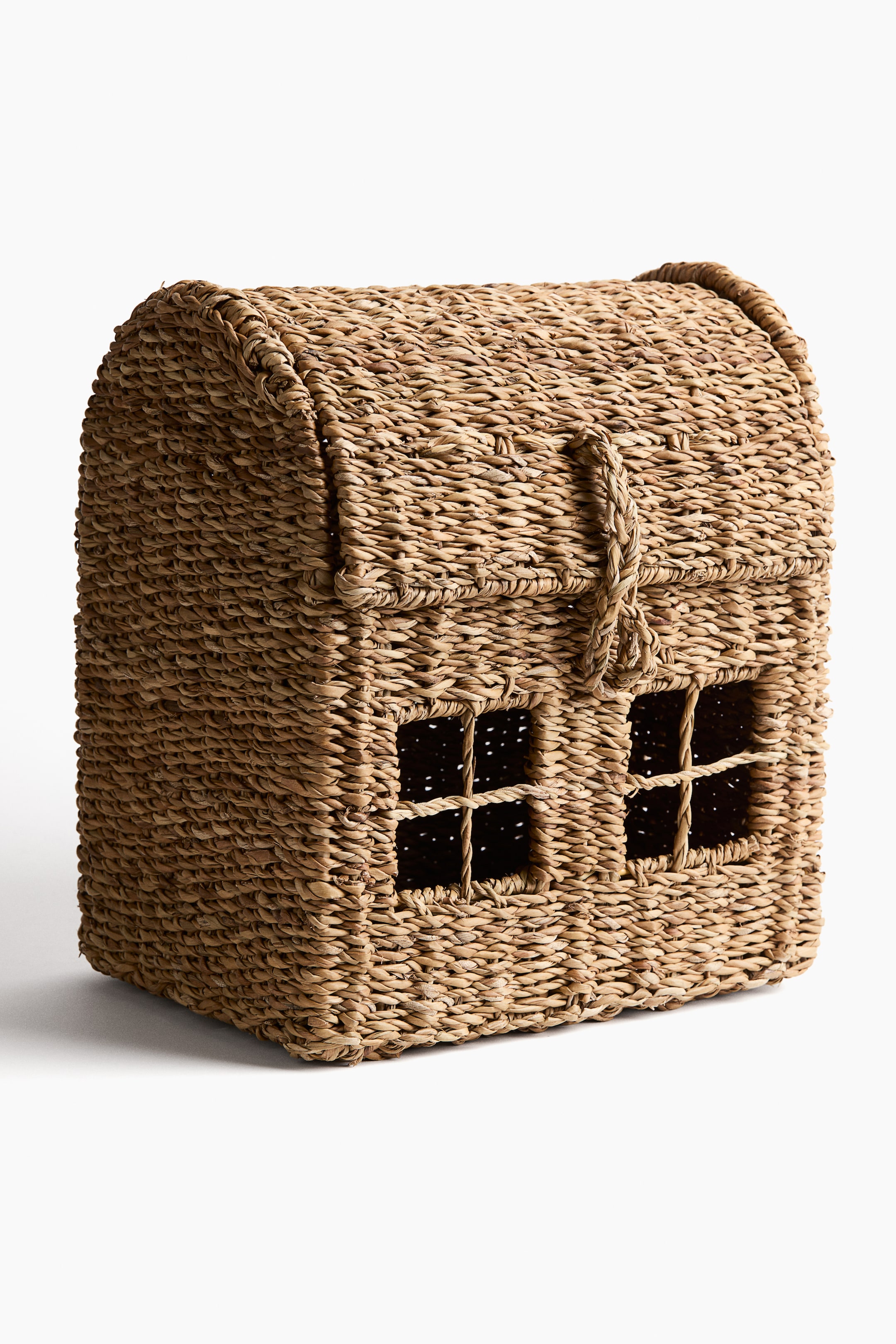 View larger image: The house-shaped basket is crafted from woven natural fibres, showing its front and side views. It features an arched roof, two rectangular window cut-outs with woven crossbars on the front, and a braided loop hanging from the roof above the windows.