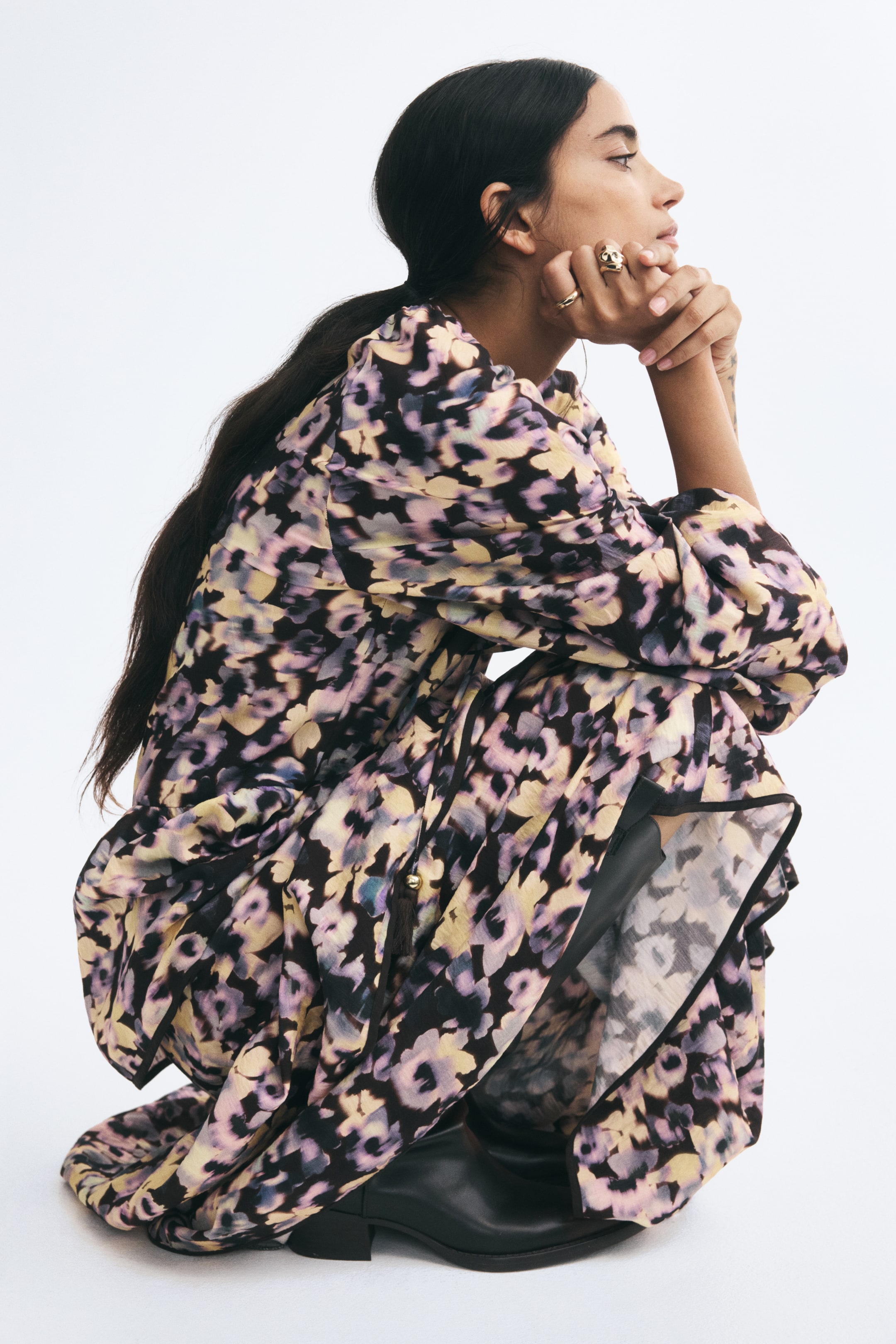 View larger image: A long dress with an all-over abstract floral print in purple, yellow, and black is being worn by the model, who is crouching in profile. The dress features long balloon sleeves and a flared, tiered skirt that is draping over a pair of black boots.