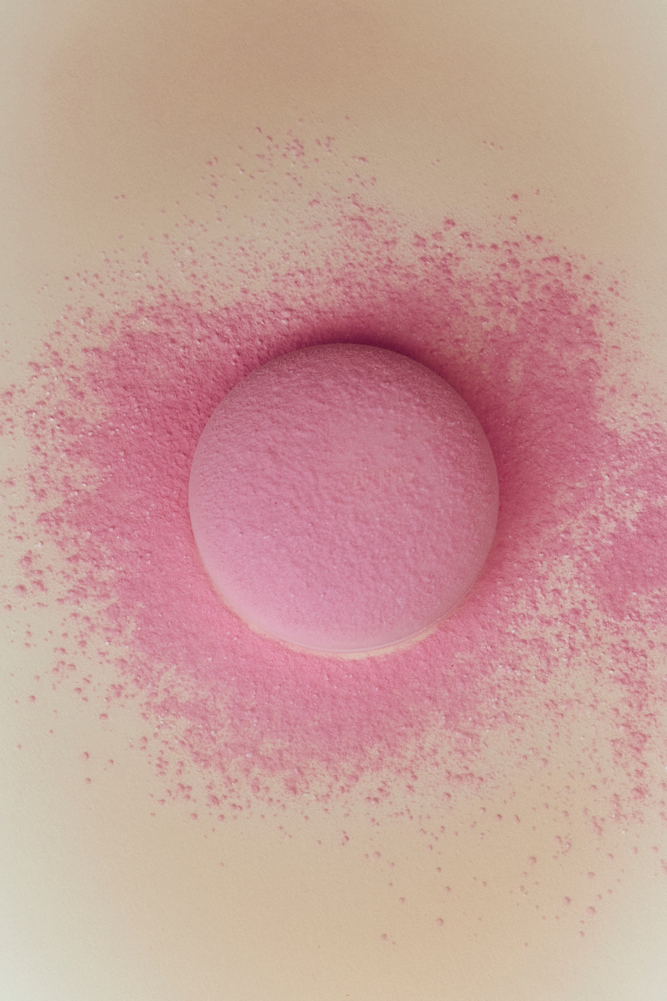 View larger image: The cream-to-powder blush appears in a round, dome shape with a pink hue. Fine pink powder is visibly scattered around its base, highlighting its powdery nature.