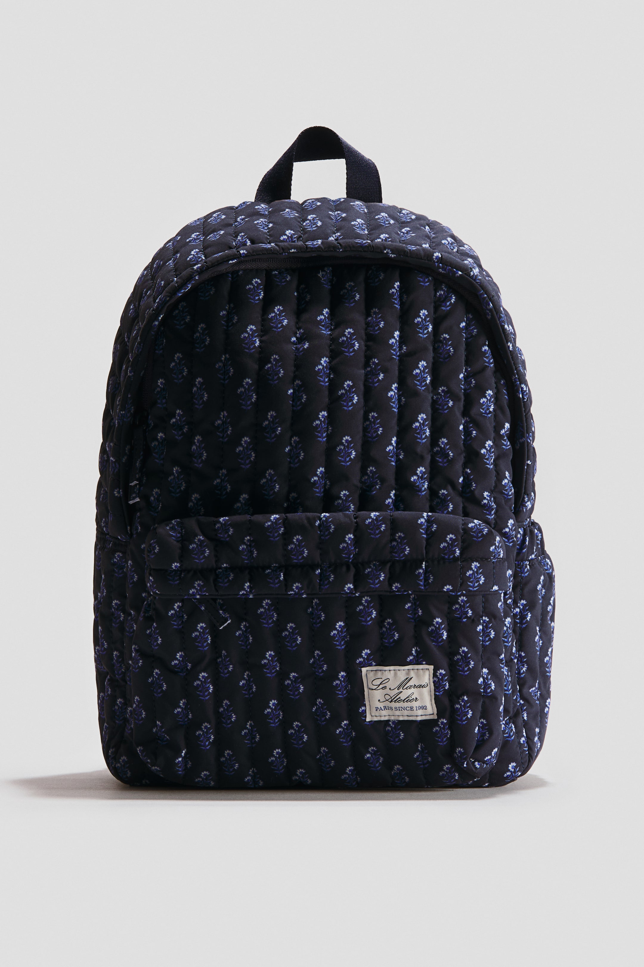 View larger image: A quilted backpack is facing front, featuring a dark blue fabric with a repeating light blue floral pattern. It is presenting a top handle, a zippered front pocket, and a rectangular brand label reading 'Le Marais Atelier PARIS SINCE 1992' attached to the bottom right of the pocket.