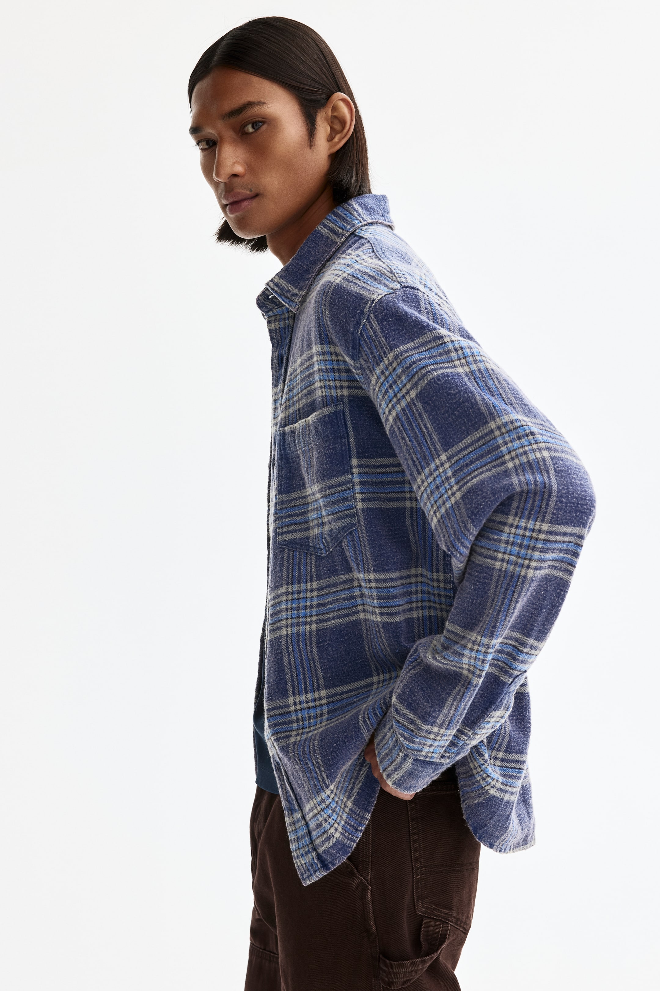 View larger image: A blue plaid shirt is shown from a side angle, worn open over another garment. It features a collared neckline, long sleeves and a single chest pocket. The brushed fabric displays a plaid pattern of varying blue and light‑grey lines, with a relaxed fit and a curved hem.