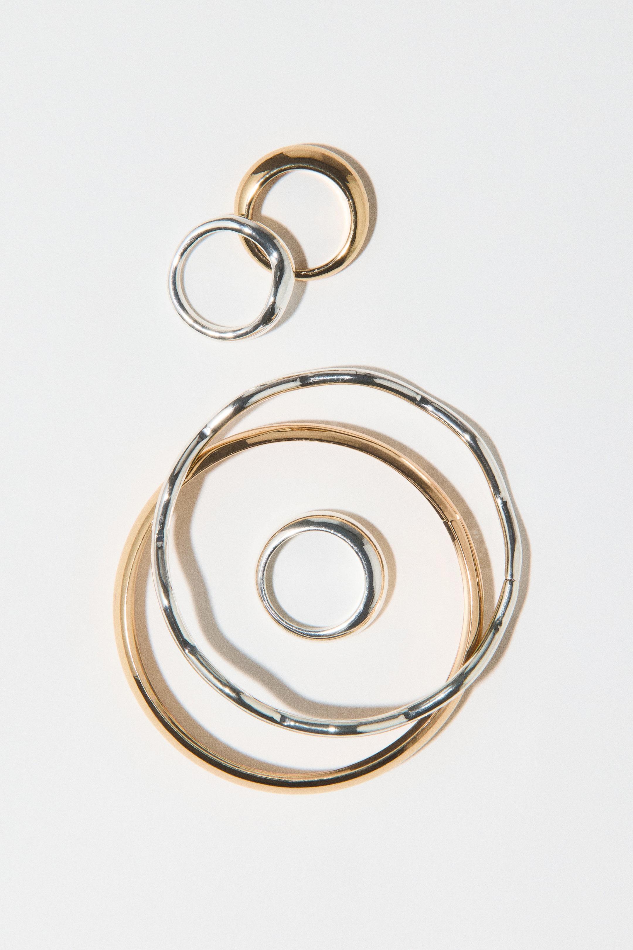 Visualizza l'immagine ingrandita: Un set di gioielli in metallo con due bracciali rigidi e tre anelli con finiture tono oro e tono argento è disposto in una vista dall’alto. Un bracciale rigido liscio tono oro e un bracciale rigido tono argento testurizzato e ondulato si sovrappongono, con un semplice anello tono argento appoggiato al centro, mentre un anello tono oro in stile dome e un anello tono argento dalla forma organica si intrecciano sopra di essi.