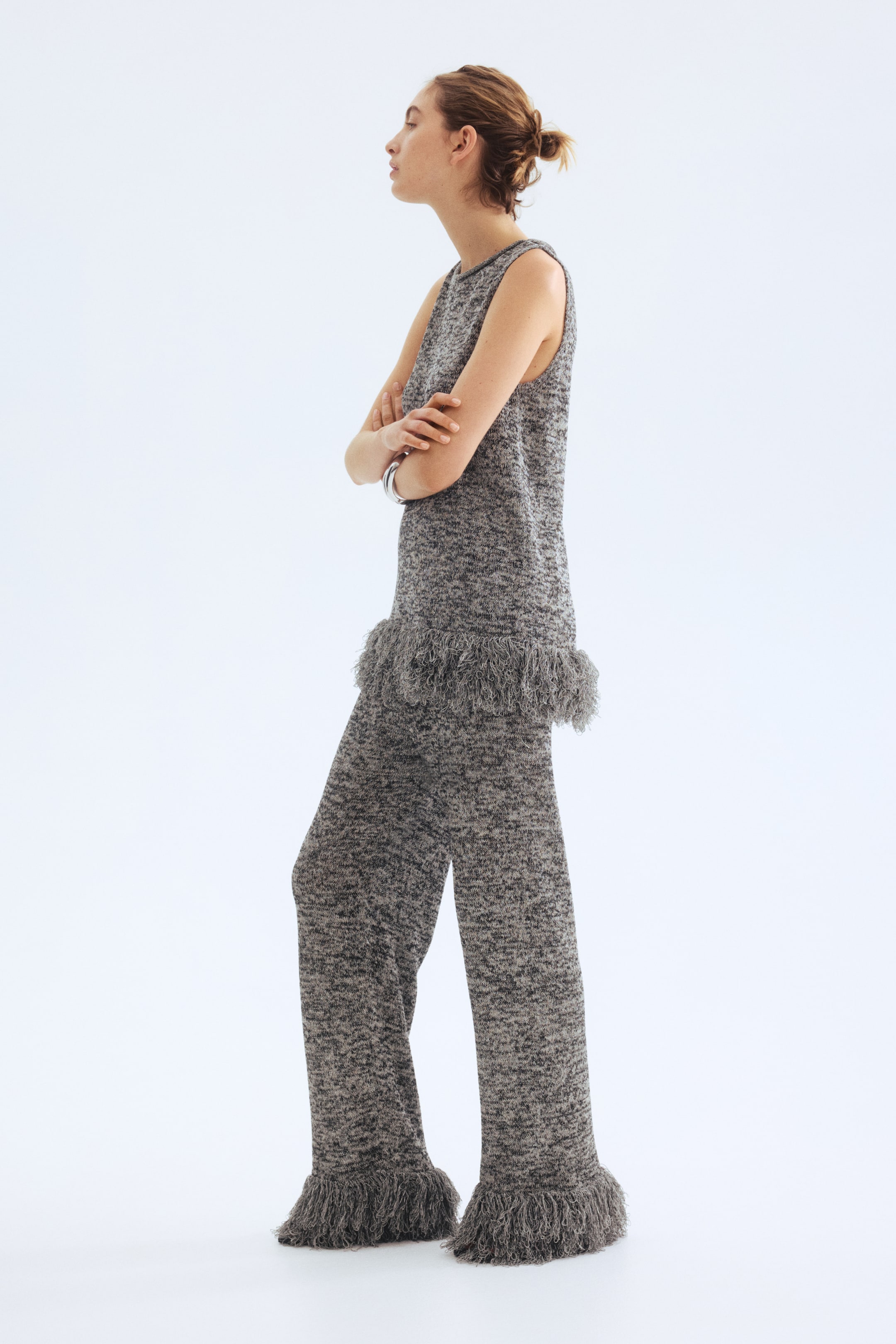 Nagyobb kép megtekintése: Egy szürke márványos kötött ujjatlan felsőt és hozzá illő bő szárú nadrágot mutatnak be oldalnézetből, jobbra nézve. Az ujjatlan felső kerek nyakkivágással és rojtos részletekkel rendelkezik a szegélyénél, míg a bő szárú nadrág rojtos részleteket mutat be a bokáknál.