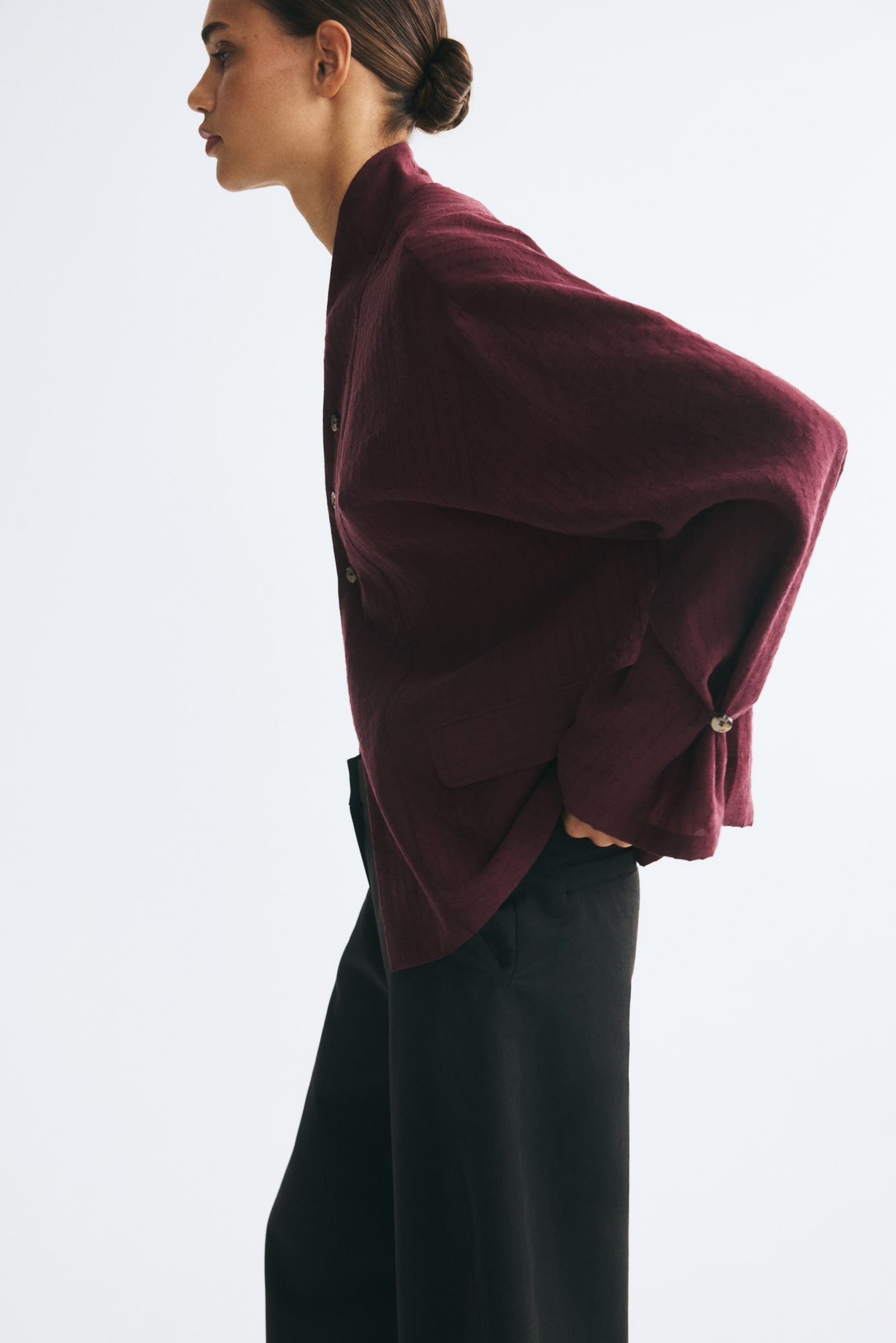 An oversized burgundy shirt in a textured weave is being worn by the model, paired with black trousers. The shirt features a stand collar, an offset front button fastening, dropped shoulders, and long sleeves detailed with button and loop fasteners. - 1