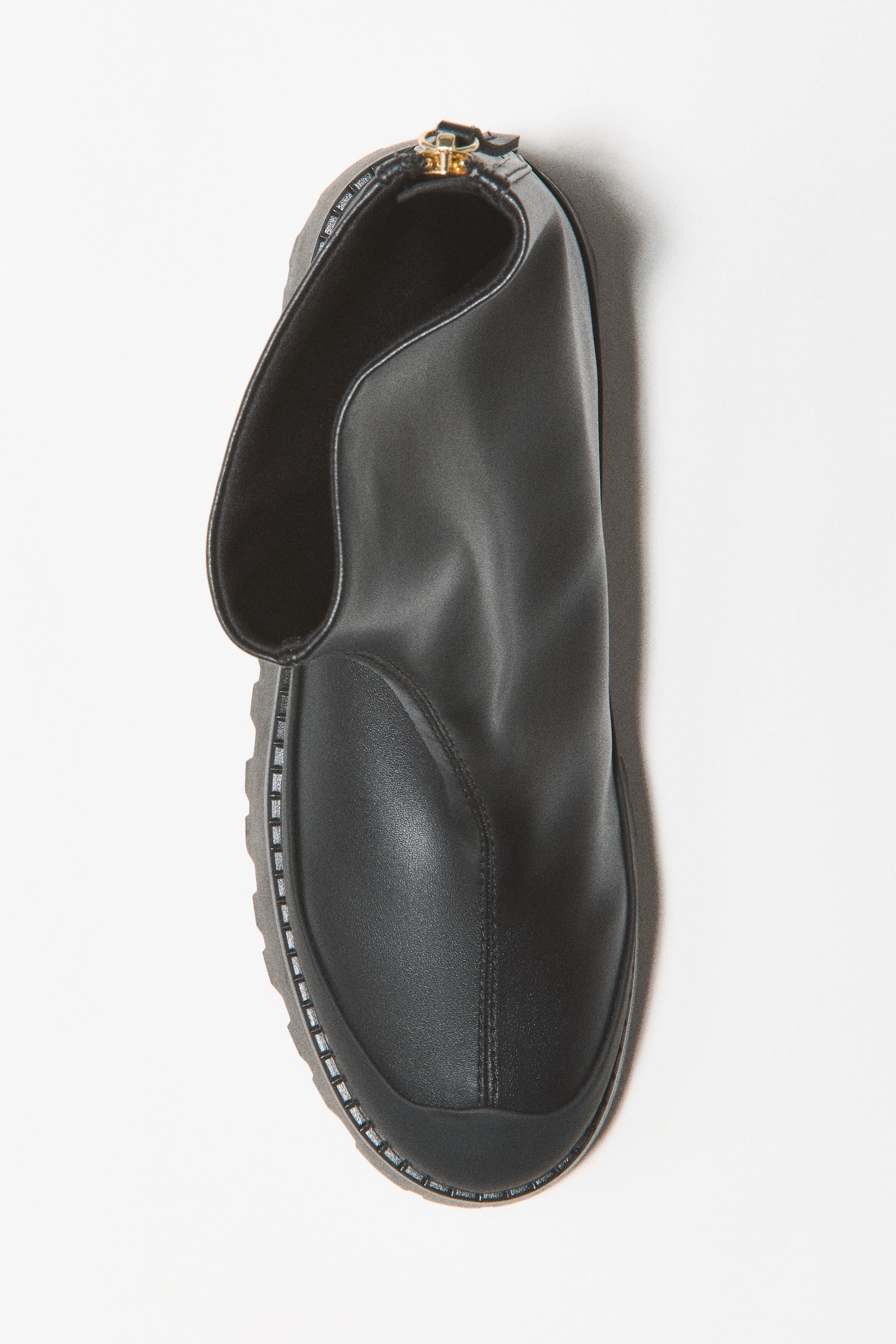 Größeres Bild ansehen: Ein schwarzer Ankle Boot ist in Draufsicht zu sehen und zeigt eine abgerundete Zehenpartie mit schützendem Mudguard sowie eine mittige Naht. Der Stiefel verfügt über einen breiten Schaftabschluss mit einem goldfarbenen Reißverschluss auf der Rückseite und eine grobe Sohle mit strukturierter Detailgestaltung.