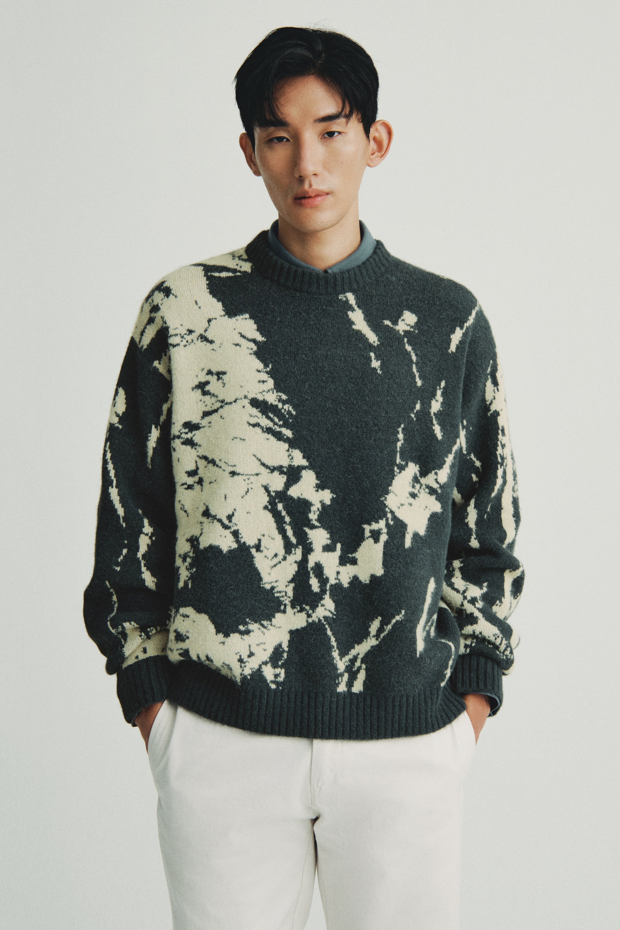 이미지 확대 보기: 크루넥 스웨터는 편안한 핏과 진한 차콜과 오프 화이트 색조의 추상적인 얼룩무늬 패턴을 보여줍니다. 리브드 커프스와 리브드 밑단이 특징이며, 네크라인 아래에는 파란색 카라 셔츠가 보입니다.