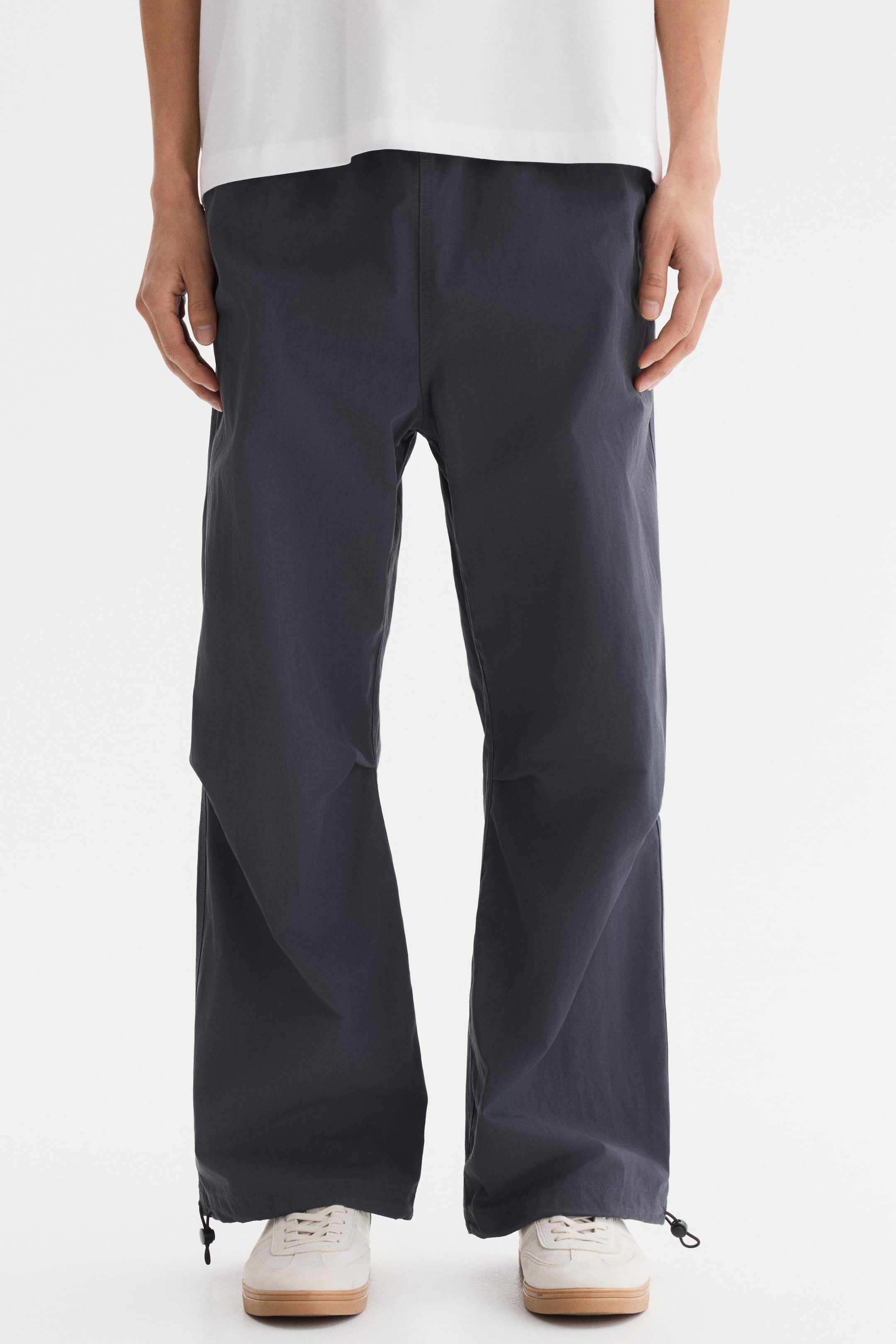 Ampliar la imagen: Pantalones de pierna ancha color gris oscuro presentados de frente, con pretina elástica y cordones ajustables en los tobillos. Los pantalones caen sueltos sobre zapatos de color claro.