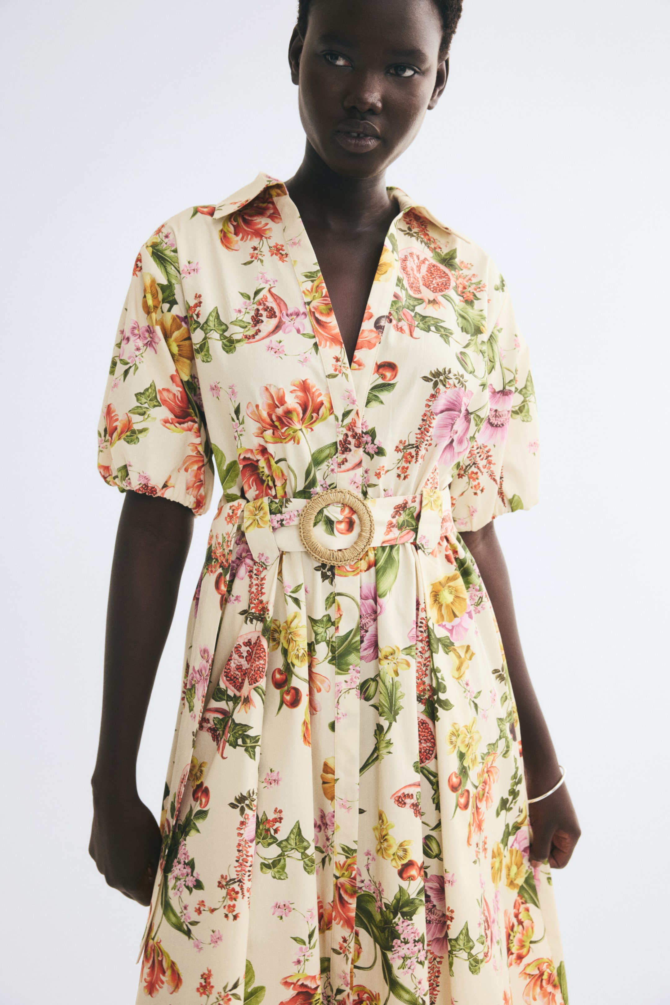 Agrandir l'image: Une robe de couleur crème ornée d’un imprimé floral et fruité éclatant, dotée d’un col ouvert et de manches courtes avec poignets élastiqués, portée par le mannequin. La robe est stylisée avec une ceinture assortie en tissu, agrémentée à la taille d’une boucle ronde tressée en paille.