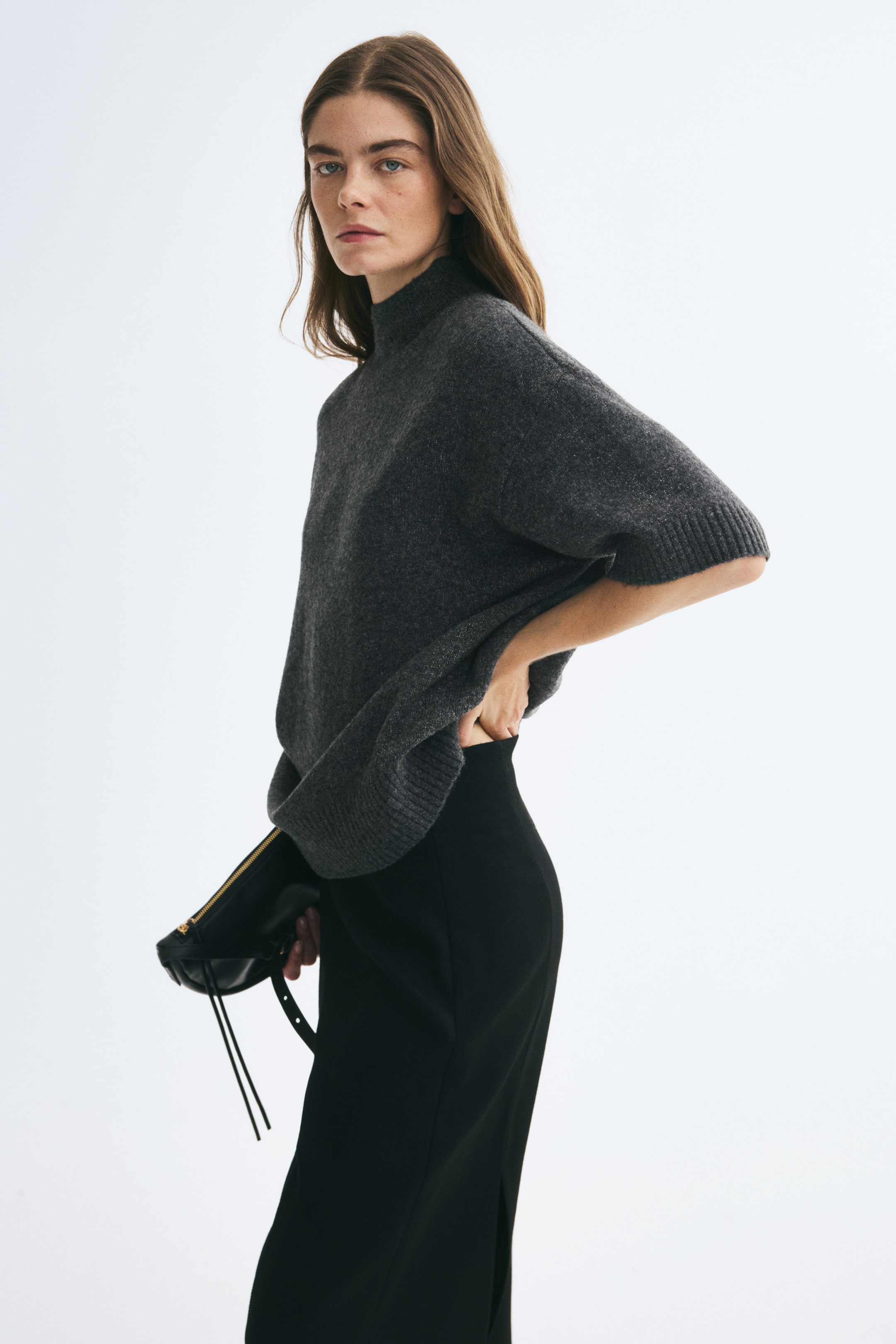 Ampliar la imagen: La modelo lleva un suéter de punto extragrande en gris oscuro, con cuello alto de canalé, hombros caídos y mangas cortas con puños de canalé. La modelo combina el suéter con una falda negra y sostiene una cartera de mano negra con cierre de cremallera.