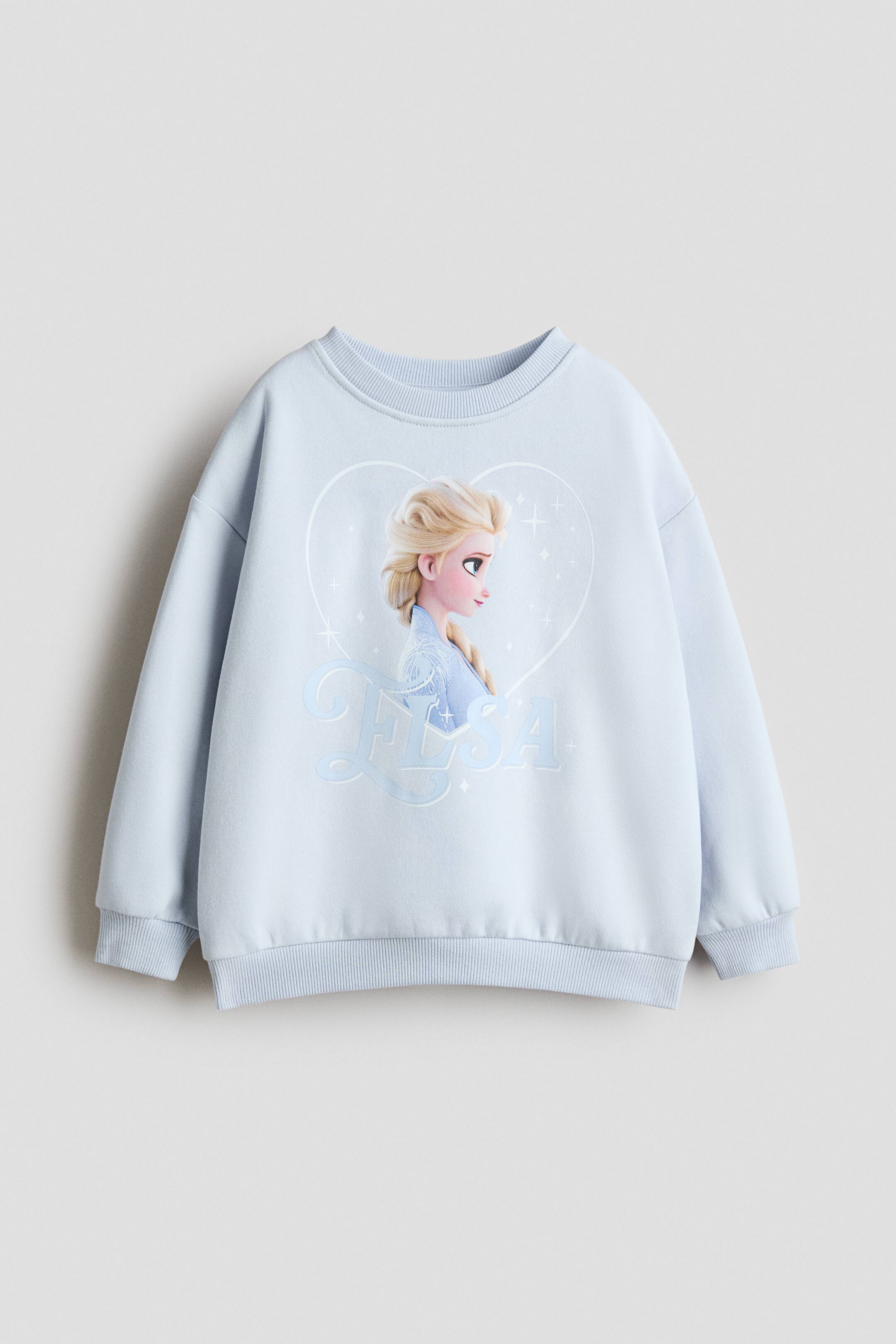 View larger image: Front view of a light blue sweatshirt in printed fabric, featuring a graphic print of a blonde-haired character in a braid looking to the right, enclosed within a subtle heart outline, with the name 'ELSA' printed below. It features a round, rib-trimmed neckline, long, voluminous sleeves, and ribbing at the cuffs and hem.