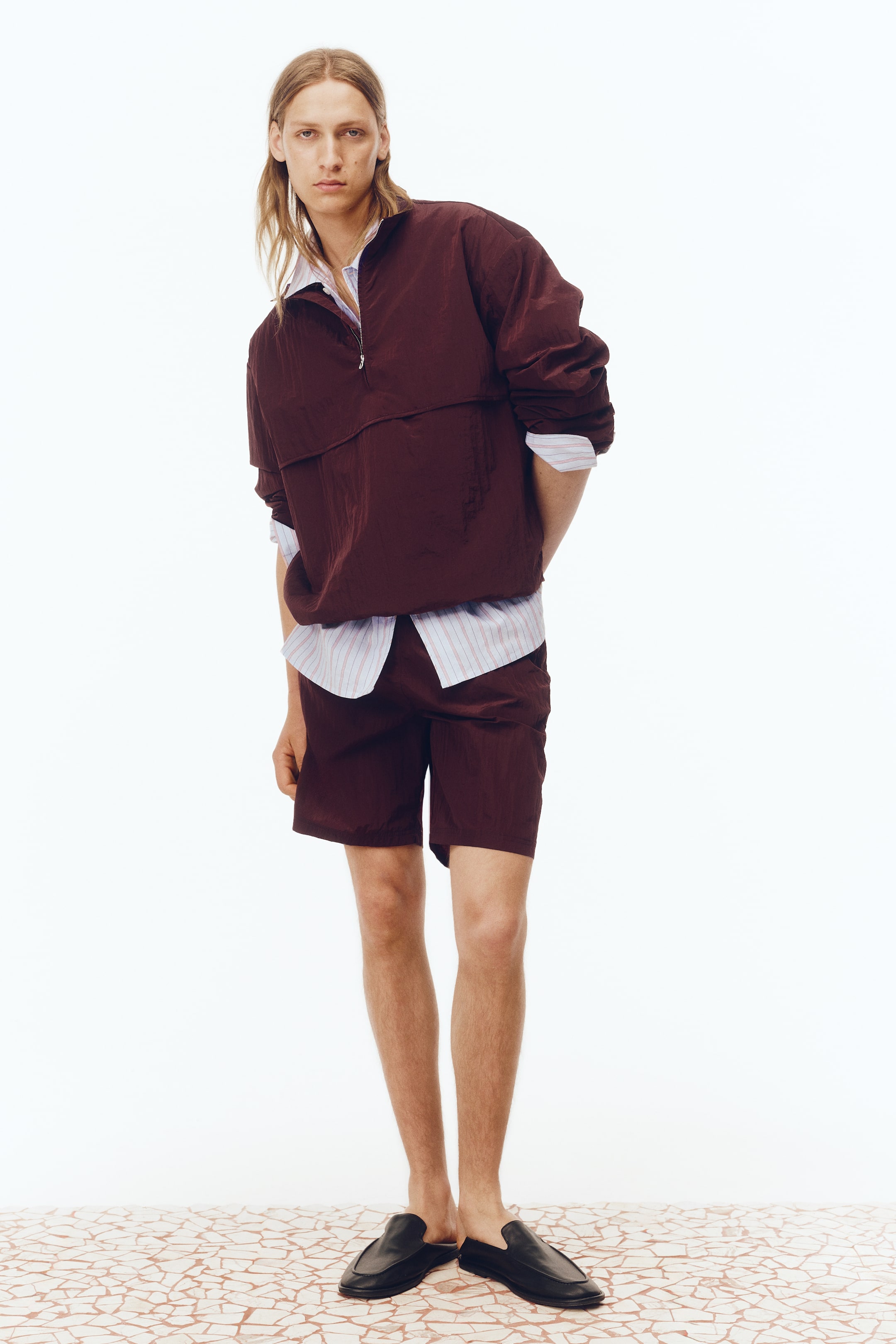 Größeres Bild ansehen: Ein bordeauxfarbener, geknitterter Windbreaker und passende Shorts werden vom Model getragen. Der Windbreaker verfügt über einen Viertelreißverschluss vorne und ist über einem gestreiften Hemd mit Kragen geschichtet.