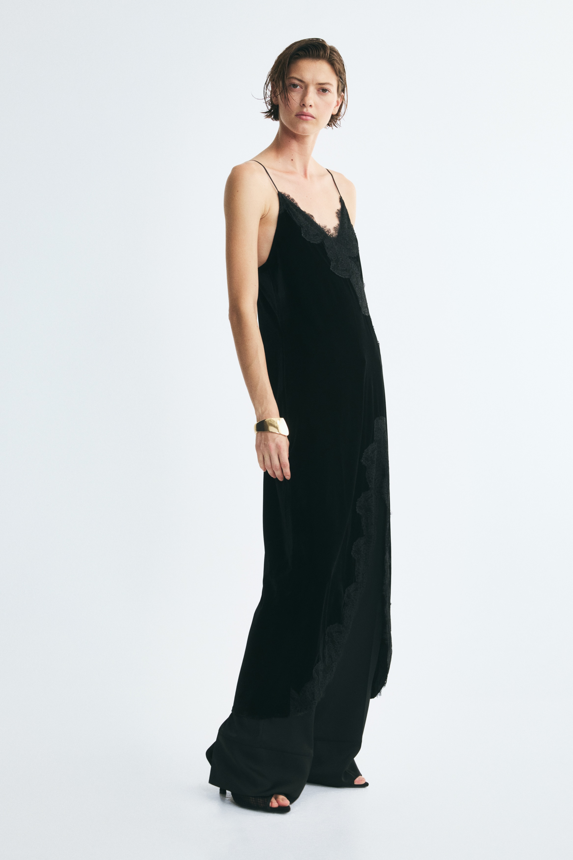 Een zwarte fluwelen slipdress wordt van voren getoond, met dunne spaghettibandjes en een V-halslijn met verfijnde kanten afwerking. De jurk toont een asymmetrische kanten applicatie op het lijfje en een hoge split aan de voorkant, afgezet met een brede, geschulpte kanten rand.