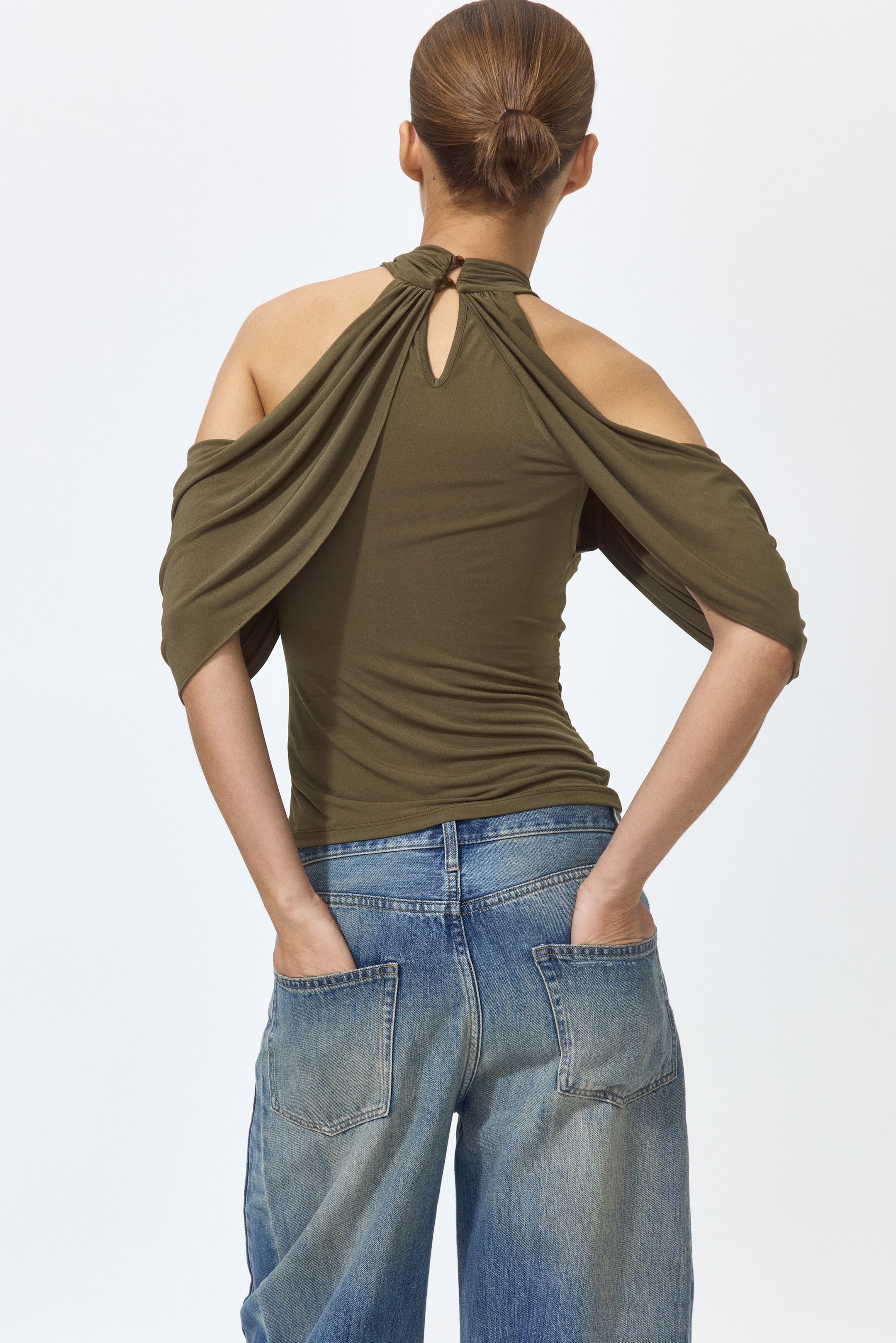 Grotere afbeelding bekijken: Een olijfgroene top wordt van achteren bekeken. Deze top heeft een hoge, gedraaide halterhalslijn met een sleutelgatuitsnede en knoopsluiting, samen met draperende off-the-shoulder mouwen en een aansluitende pasvorm.