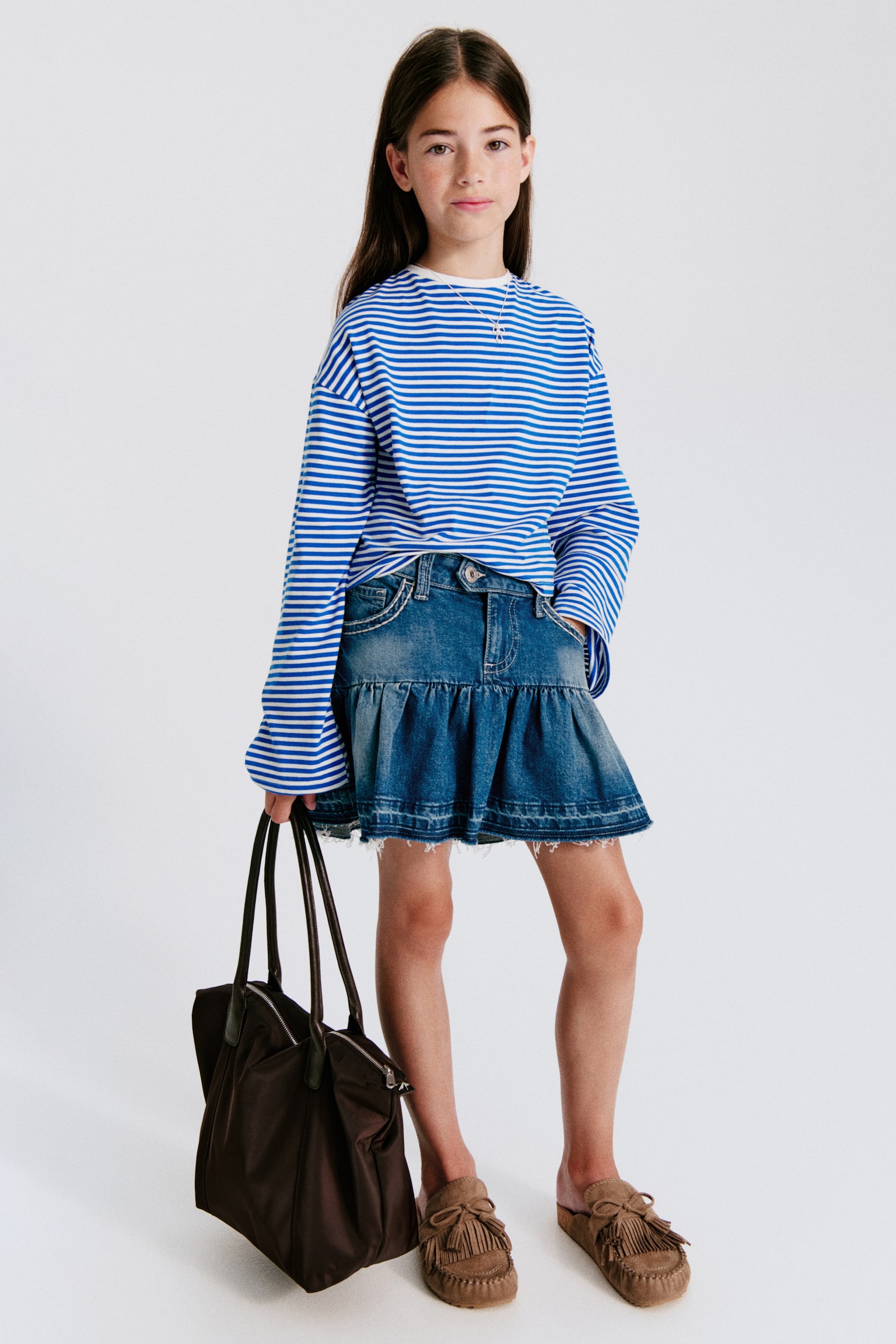 A blue denim skirt is positioned directly facing forward, displaying a medium wash with lighter fading. It comes with a button closure, zip fly, belt loops, and two front pockets featuring white contrast topstitching. A gathered ruffle tier forms the lower section, finishing in a distressed, frayed hem. - 1