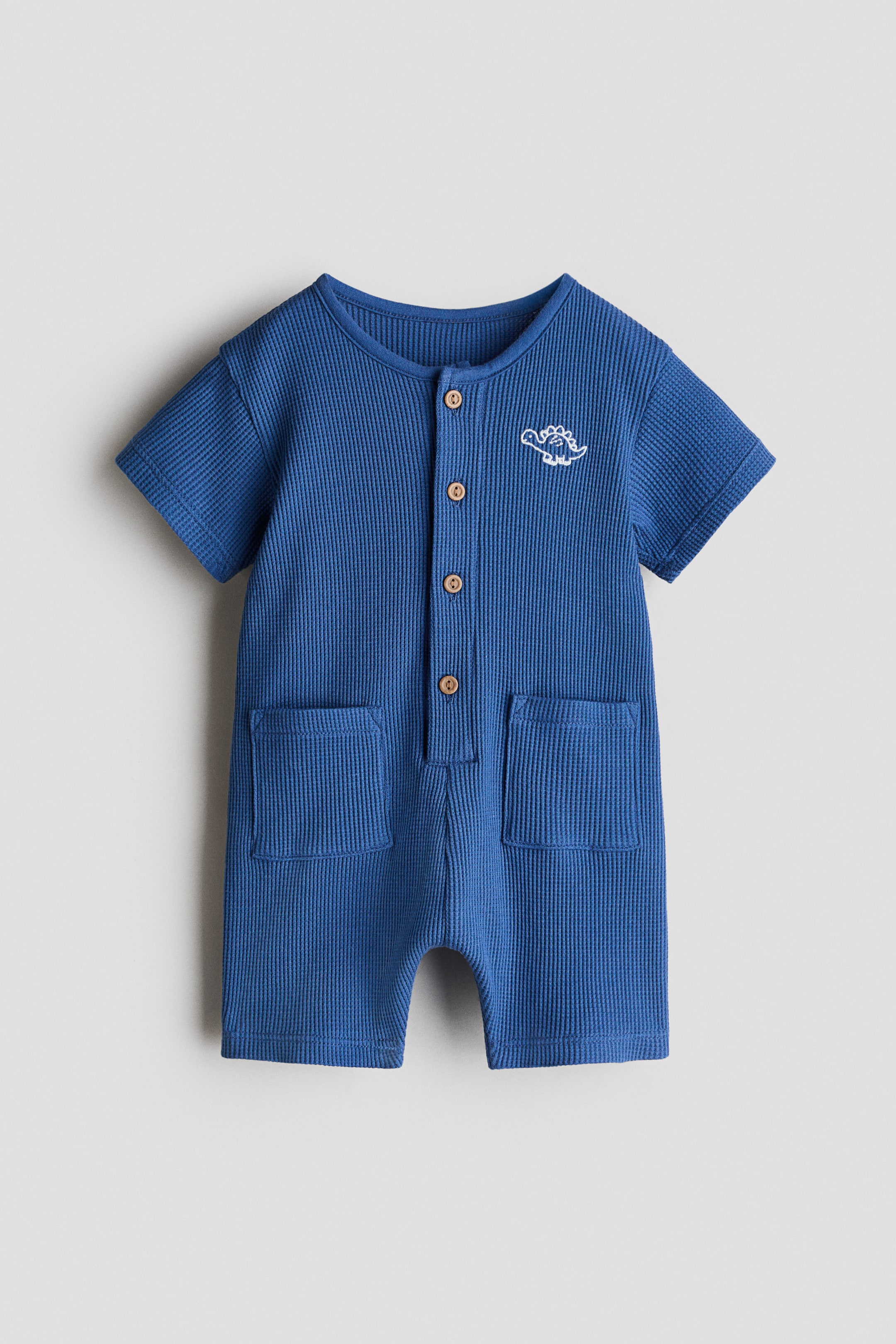 View larger image: A short-sleeved romper suit in blue waffled cotton jersey is on display. It features a round neckline, a row of wooden buttons down the front, a small white embroidered dinosaur detail on the left chest, two open patch pockets on the front, and short legs.