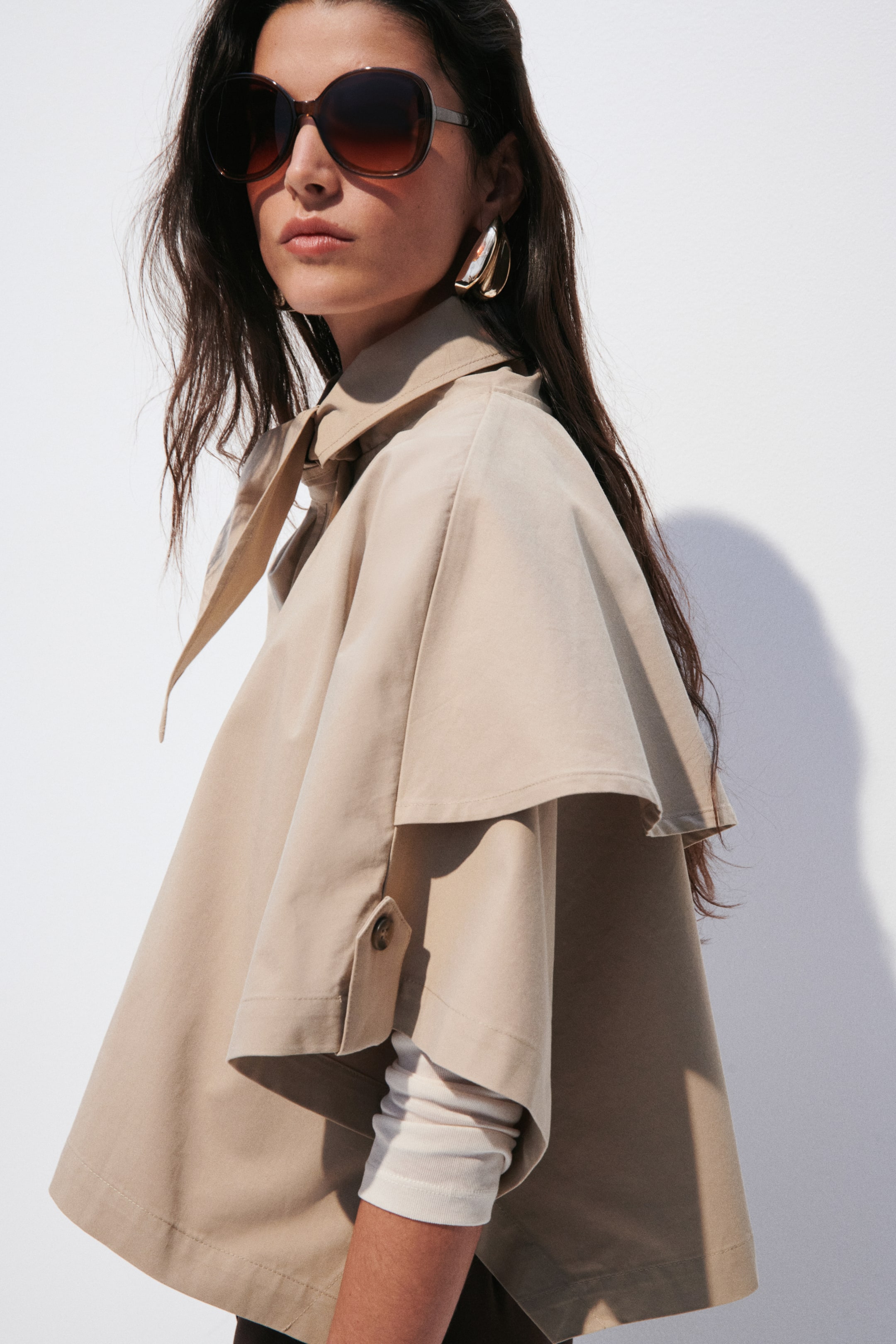 Größeres Bild ansehen: Eine beige Cape-Jacke präsentiert einen gestuften Sturmklappenüberwurf über den Schultern und weite Ärmel mit Knopfverschlüssen, während ein Bindeband eine Schleife am Stehkragen bildet. Ein weißes, langärmliges Oberteil ist unter den weiten Ärmeln sichtbar.