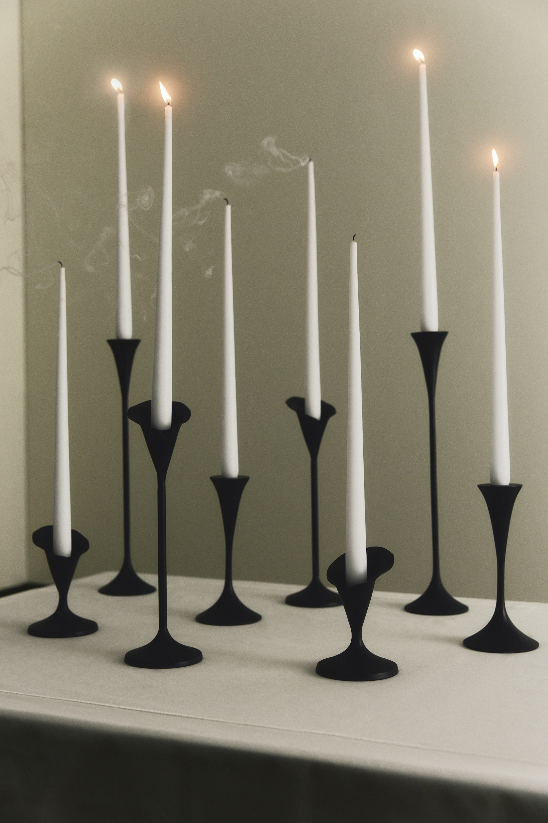 Voir une image plus grande: Un groupe de chandeliers en métal noir mat de hauteurs variées est disposé ensemble, chacun présentant une douille en forme de fleur, une tige élancée et une base large et évasée. Les chandeliers soutiennent de hautes bougies chandelles blanches, dont certaines sont allumées avec des flammes actives et d’autres dégagent de fines volutes de fumée.