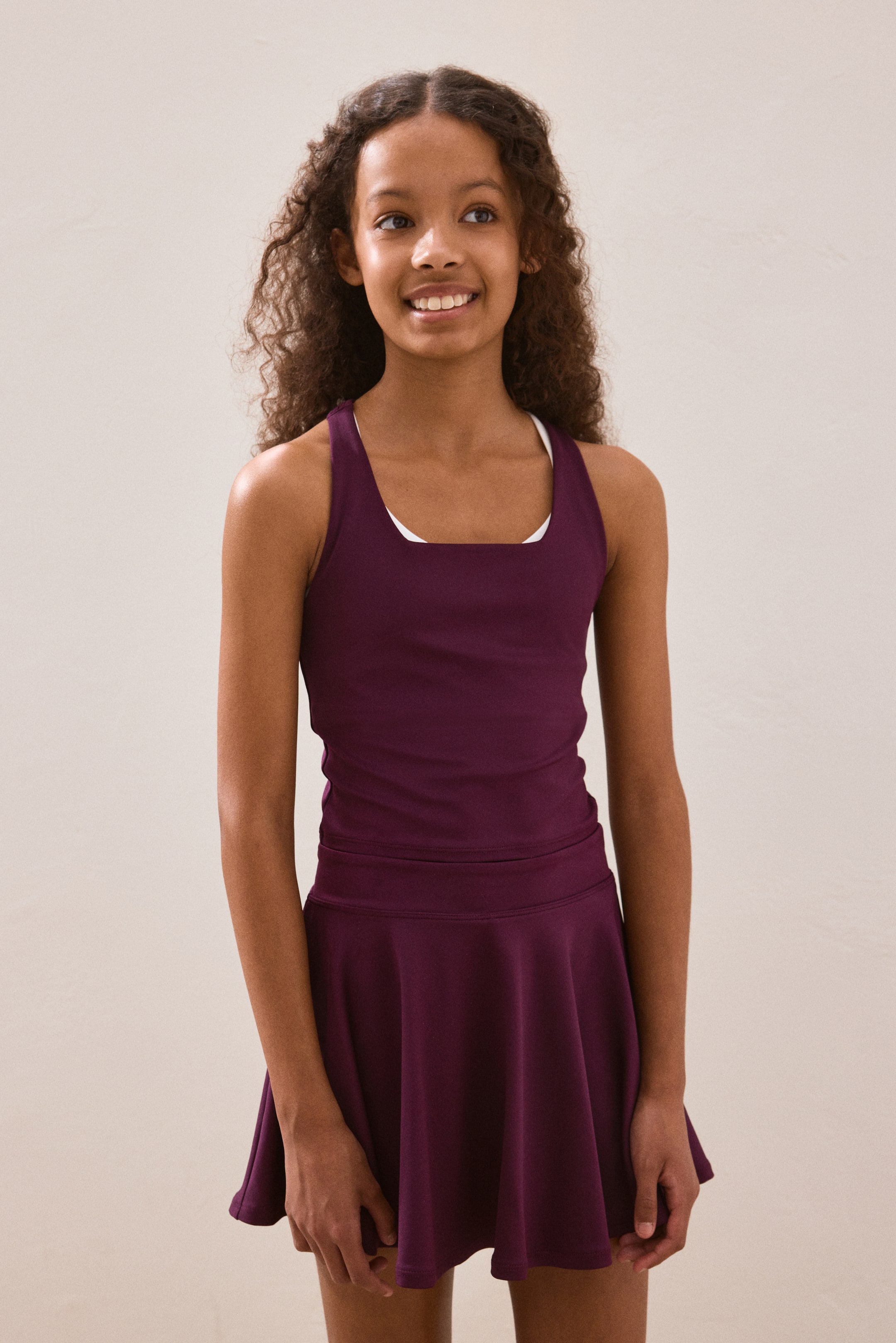 Grösseres Bild ansehen: Ein tiefviolettes Sportset, bestehend aus einem Tanktop mit quadratischem Ausschnitt und Racerback-Design, das über einem weißen Top getragen wird, sowie einem passenden ausgestellten Rock mit breitem Bund, wird von vorne präsentiert.