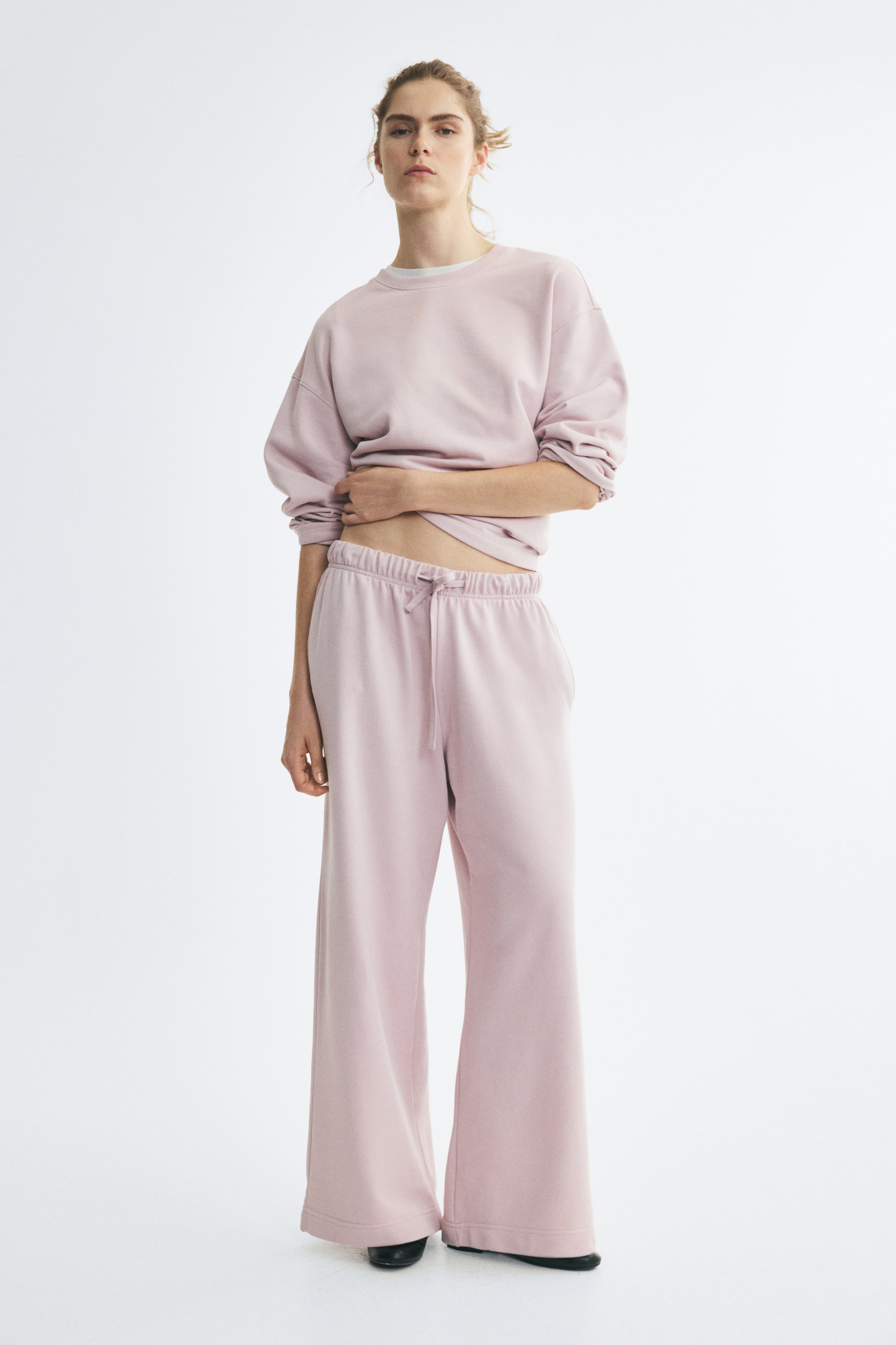 View larger image: The model is wearing light pink track pants featuring an elasticated, drawstring waist and straight, wide legs. She is pairing the pants with a matching long-sleeved sweatshirt, lifting the hem slightly with one hand to show the waistband detail.