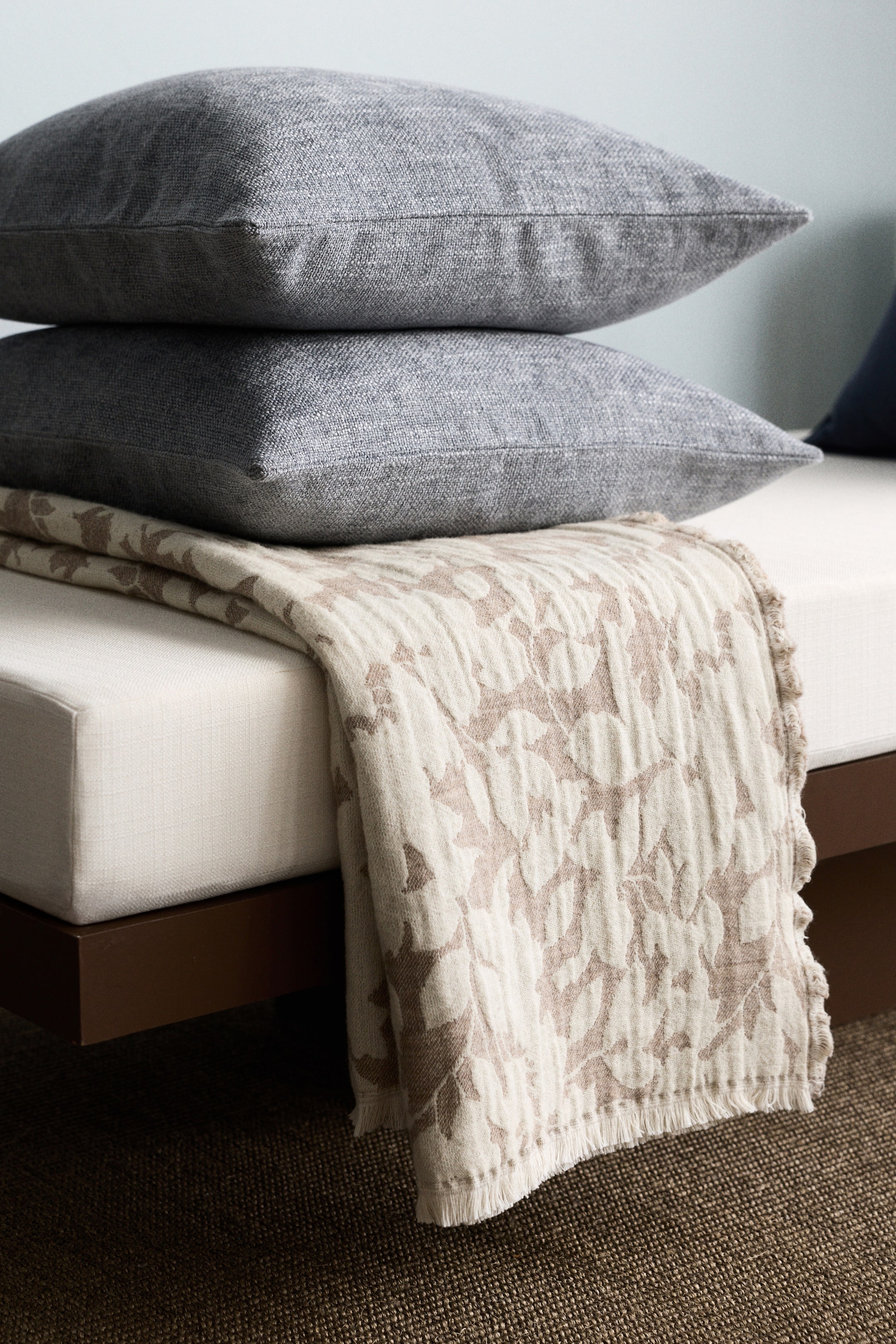View larger image: A cream-coloured blanket, featuring a textured light brown botanical pattern, is draped over a light-coloured cushion. Its fringed edge is hanging down, and two stacked grey pillows are resting on its surface.