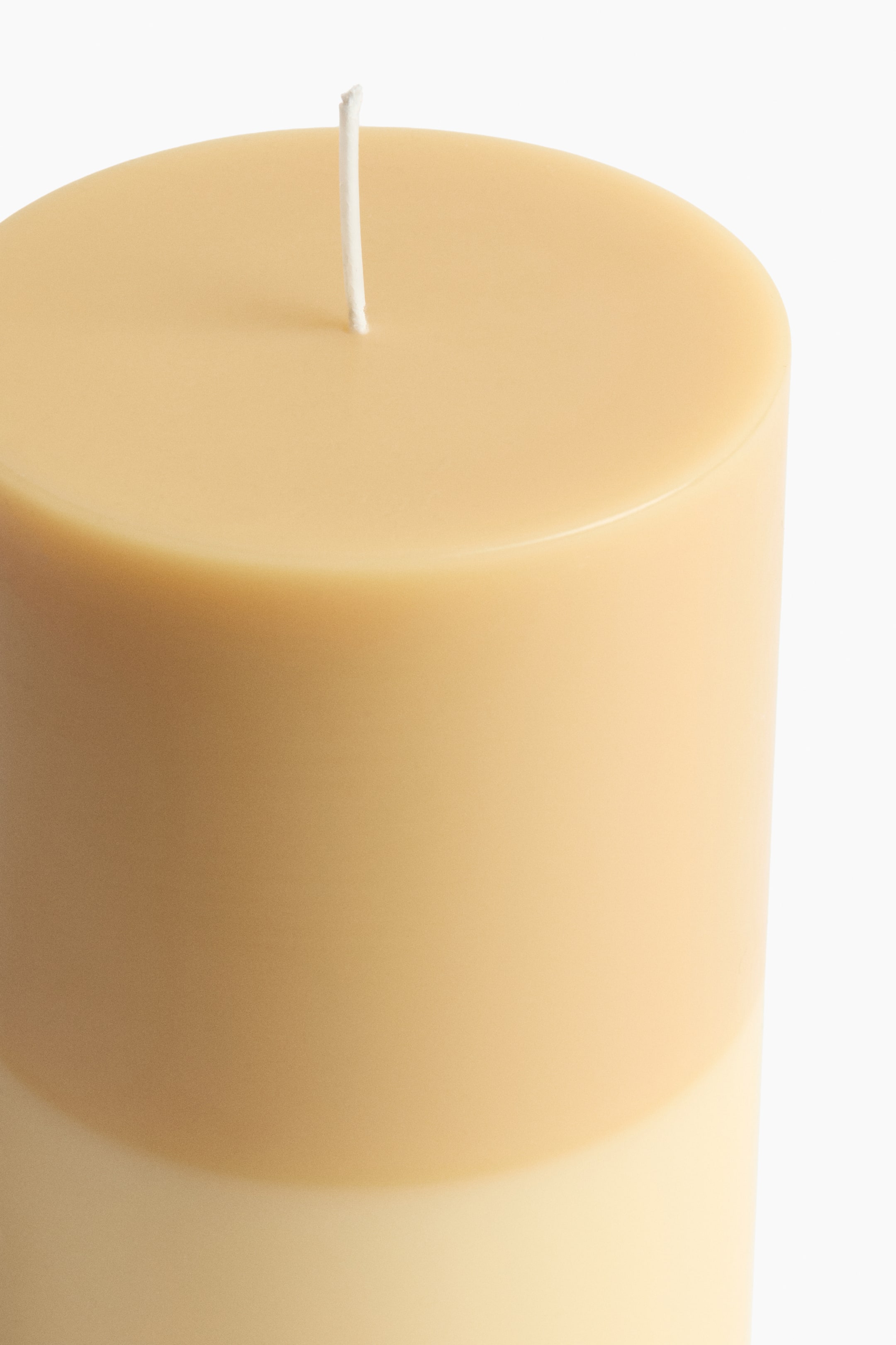 View larger image: A large pillar candle is displaying a two-tone design, with a pale tan upper section transitioning to a lighter cream on the lower part. A white wick is standing upright from its flat top surface.